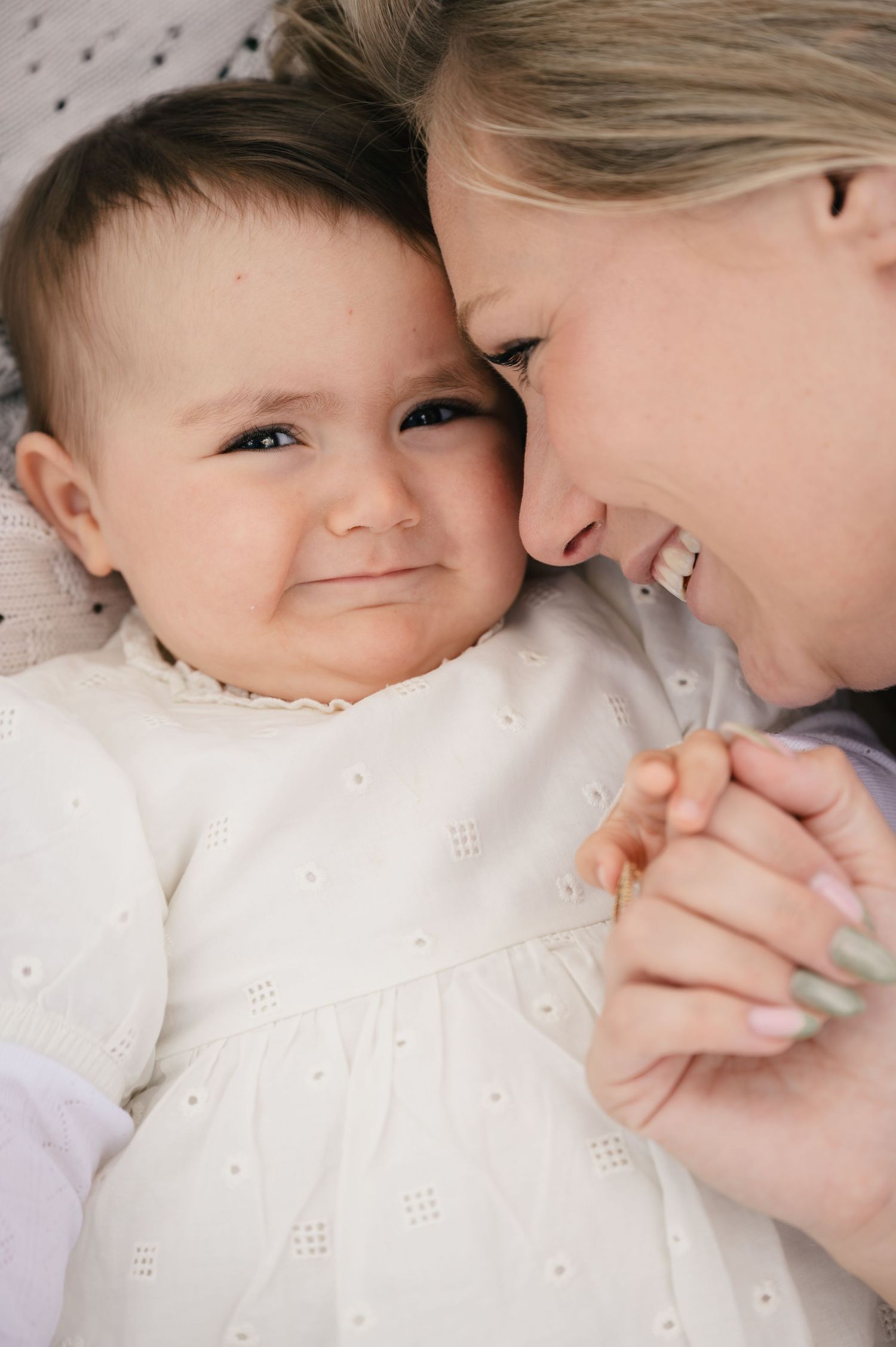 A smiling mother tenderly touches foreheads with her happy baby girl in a white dress while holding hands.