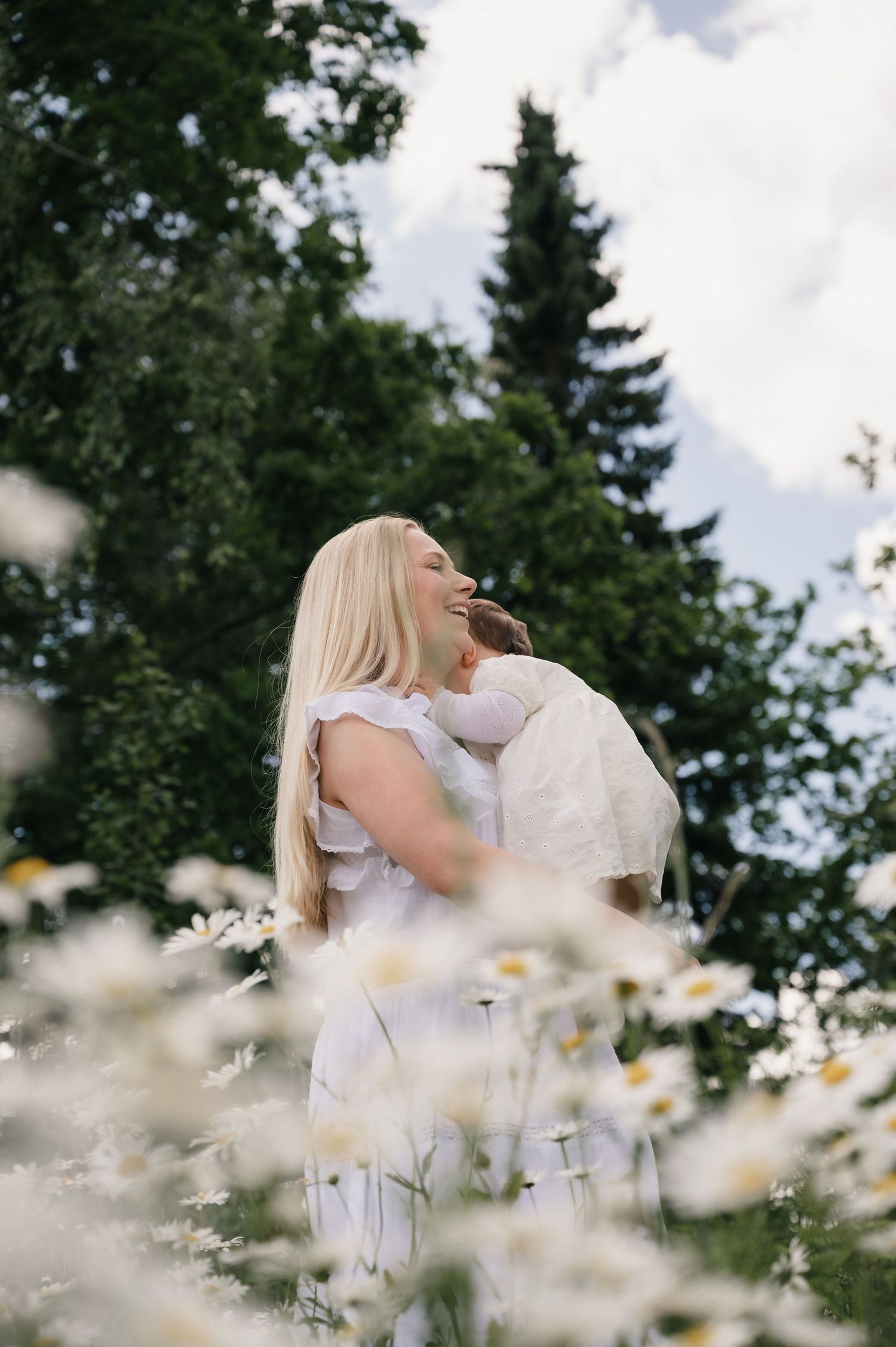 Woman in white dress holding child in daisy field with trees in background on cloudy day.