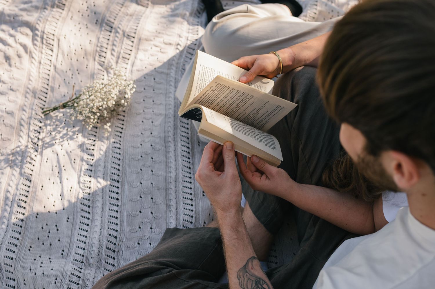 Man with tattoos reading a book while relaxing on a white blanket outdoors with dried flowers nearby.