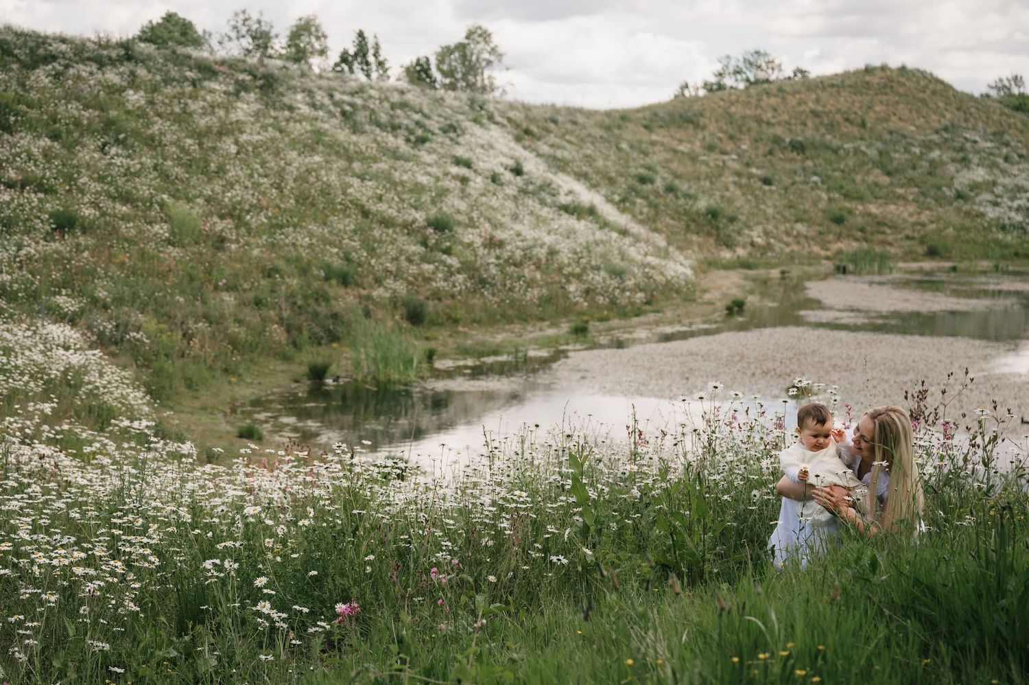 Family with baby standing in wildflower meadow near a serene pond surrounded by green hills.