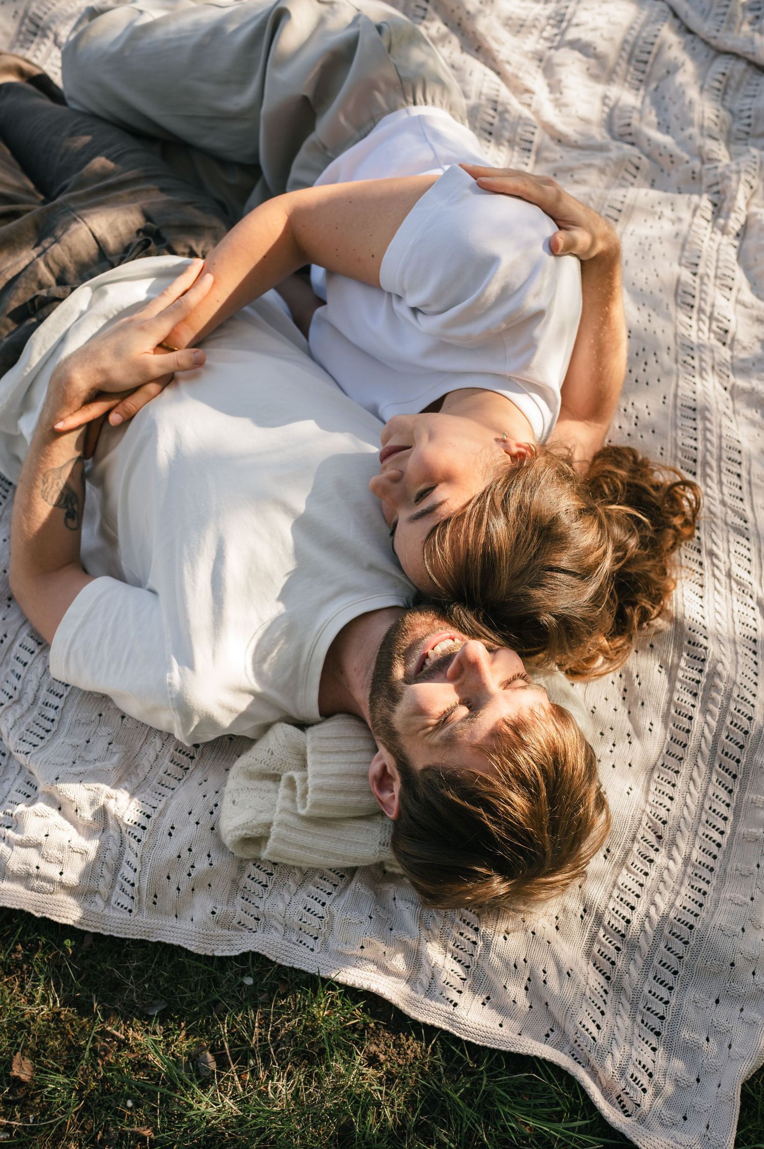 Couple lying on a knit blanket outdoors, laughing and embracing in warm sunlight during a romantic picnic.