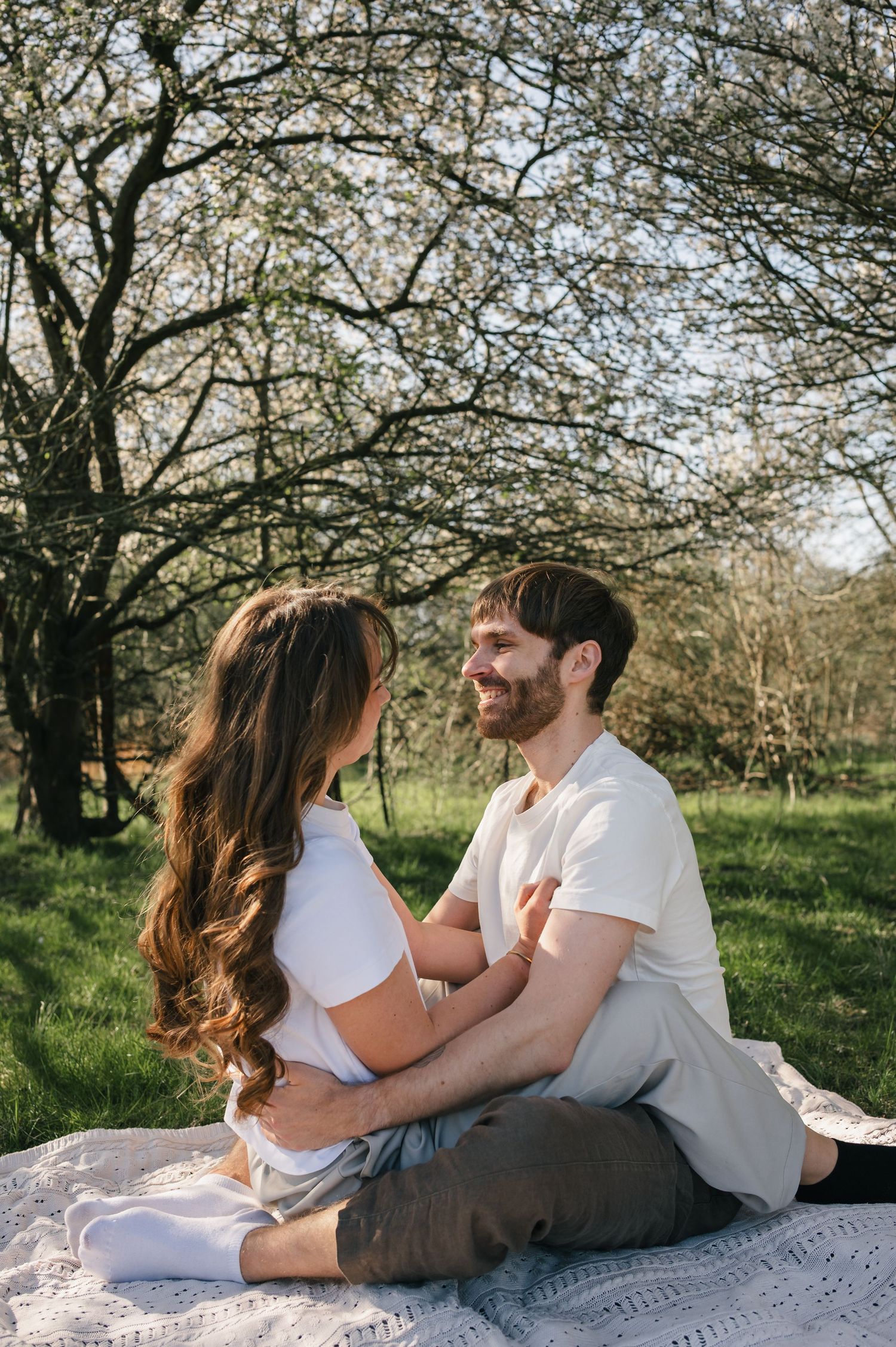 Couple sitting on a blanket in a sunny park, facing each other and smiling, with blooming trees in the background.