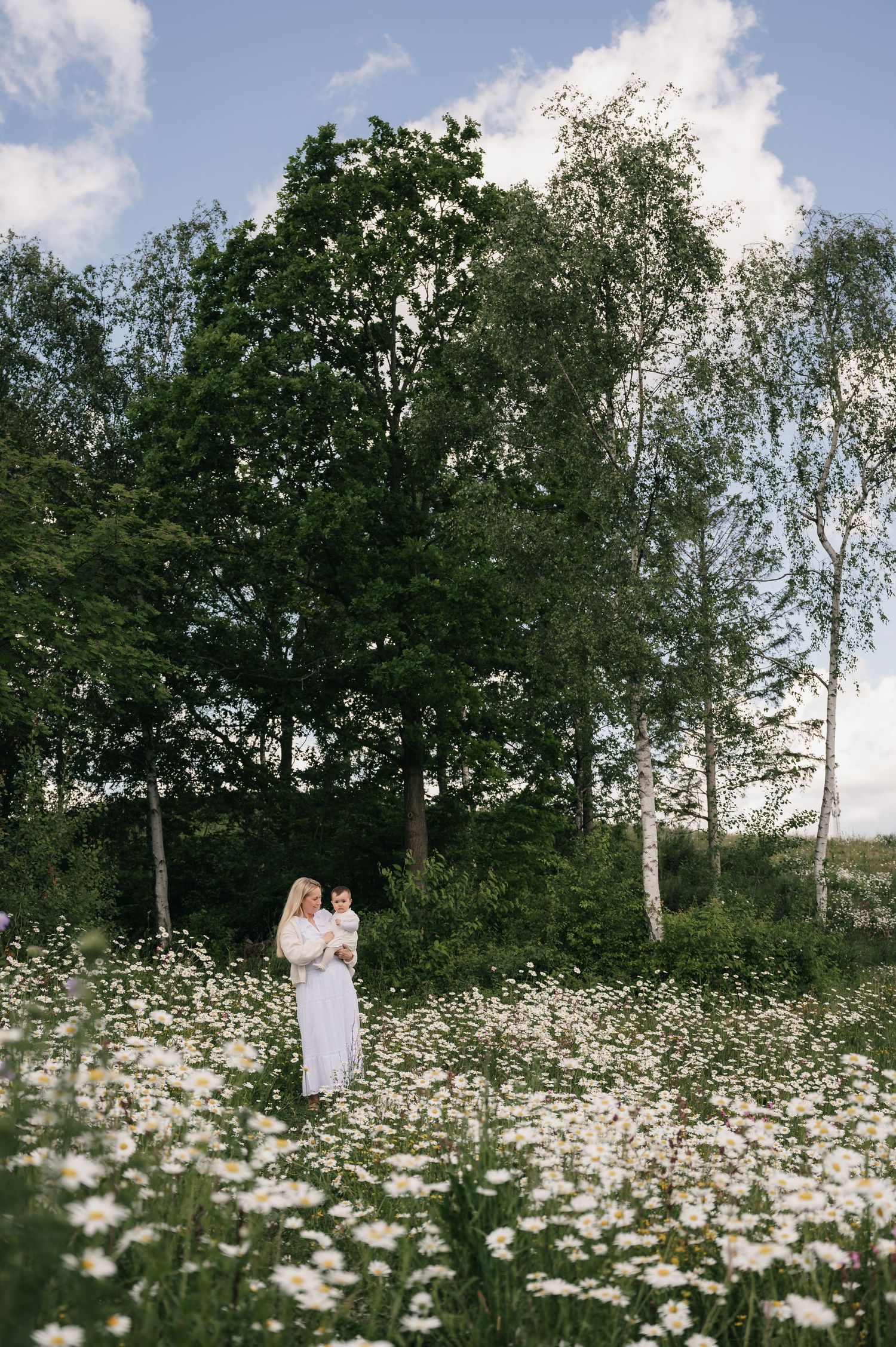 A woman in white dress holds a baby in a daisy field with lush green trees under a blue sky.