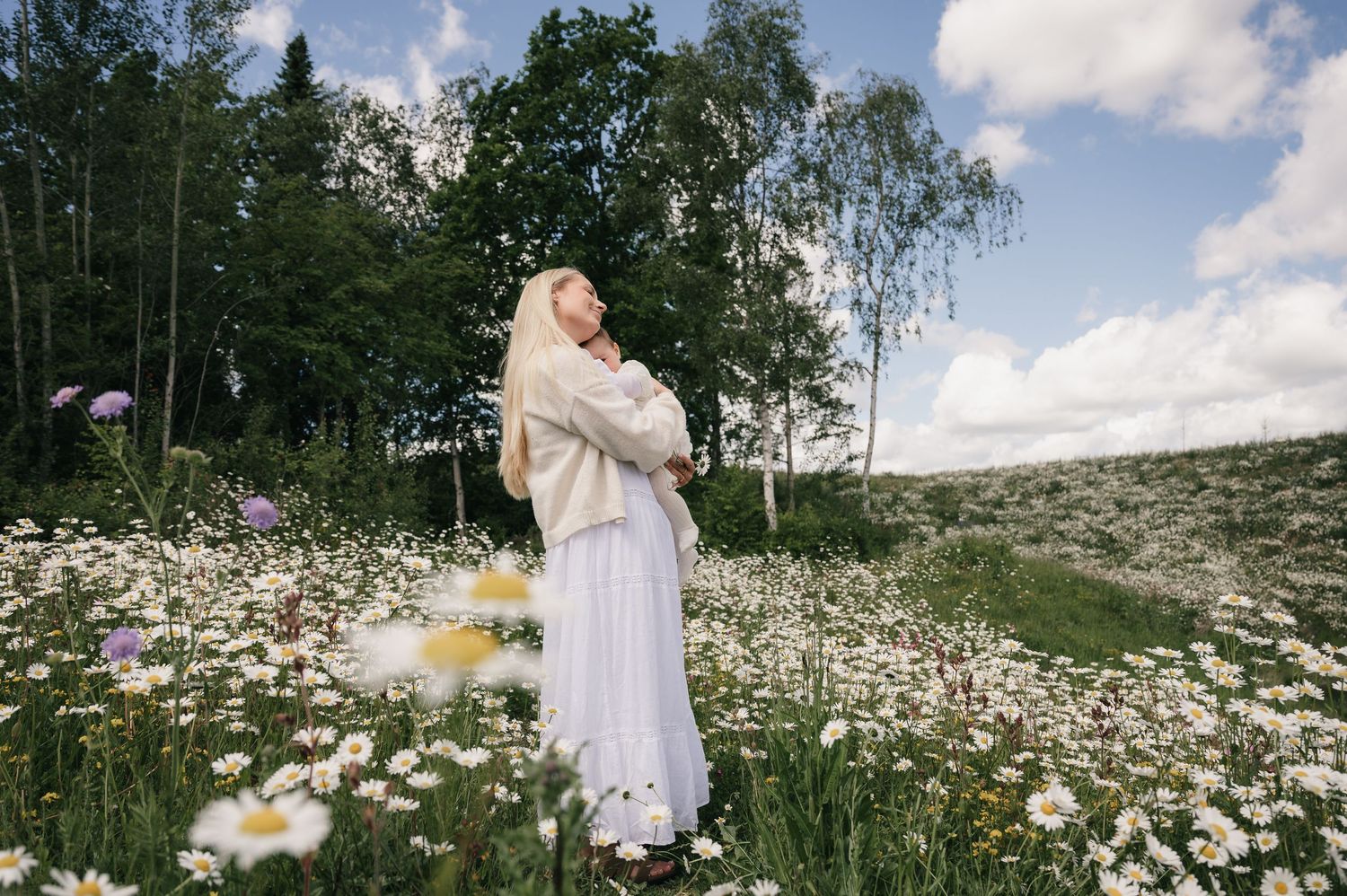 Woman in white dress and shawl standing in a field of daisies with birch trees and cloudy sky.