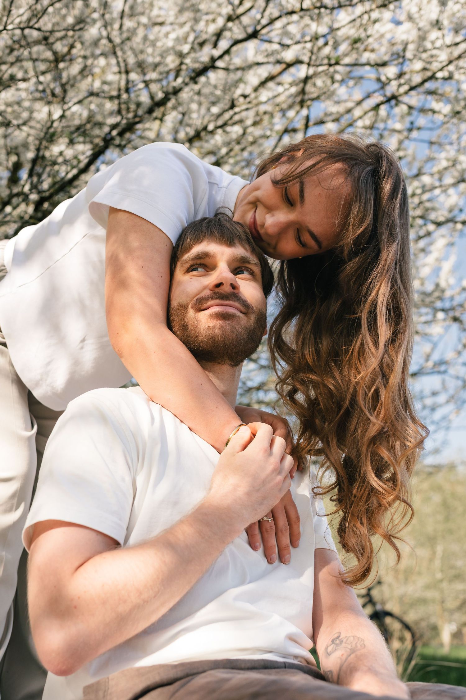 A couple embraces outdoors near blooming white trees, woman leaning over man's shoulder smiling on a sunny day.