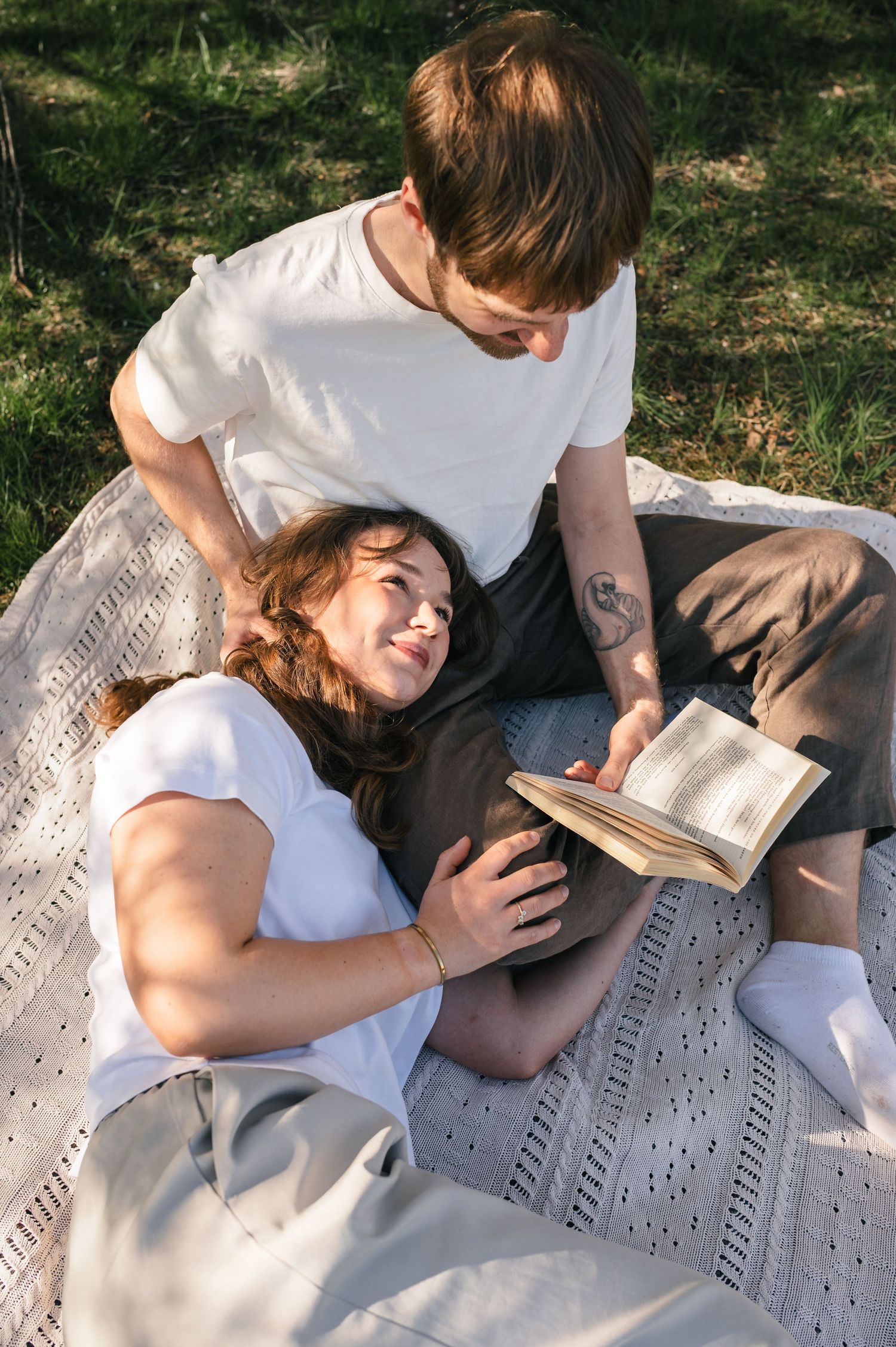 Couple relaxing on a blanket in a sunny park, woman lying on man's lap while he reads a book outdoors.