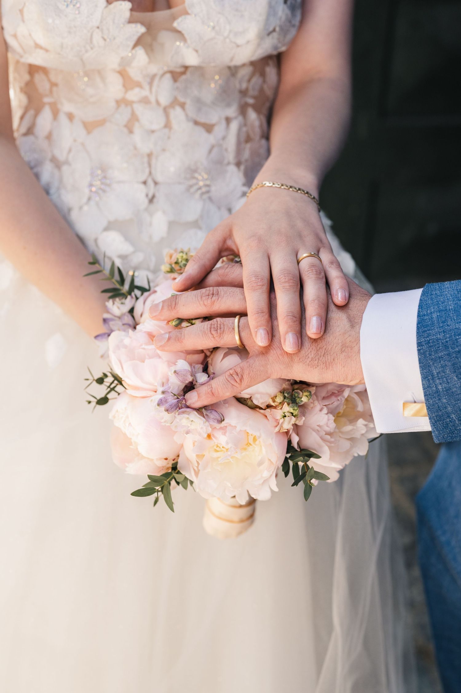Bride and groom hands with wedding rings resting on a soft pink peony bridal bouquet.