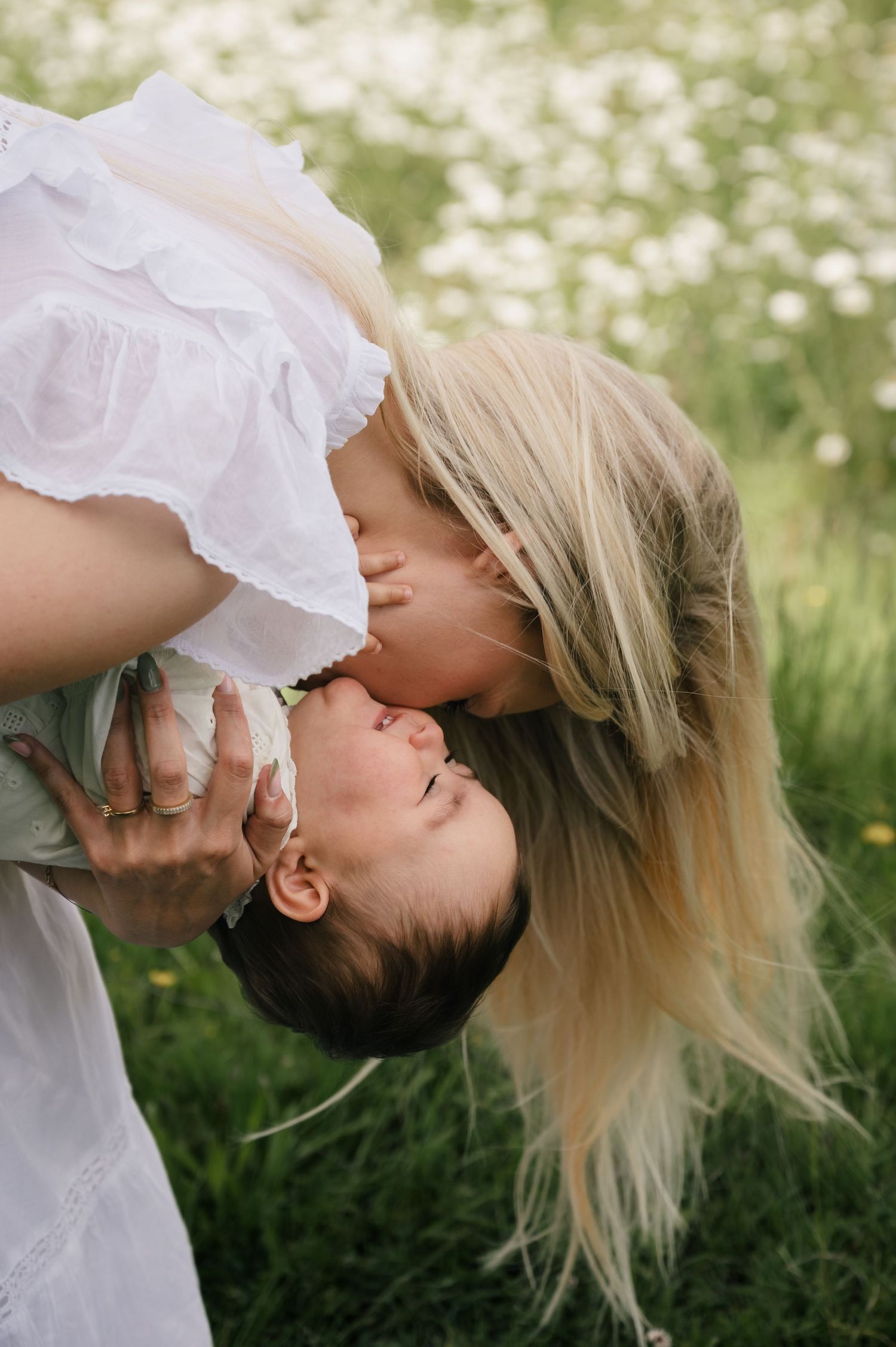 A blonde mother tenderly kisses her baby outdoors in a blooming wildflower meadow.