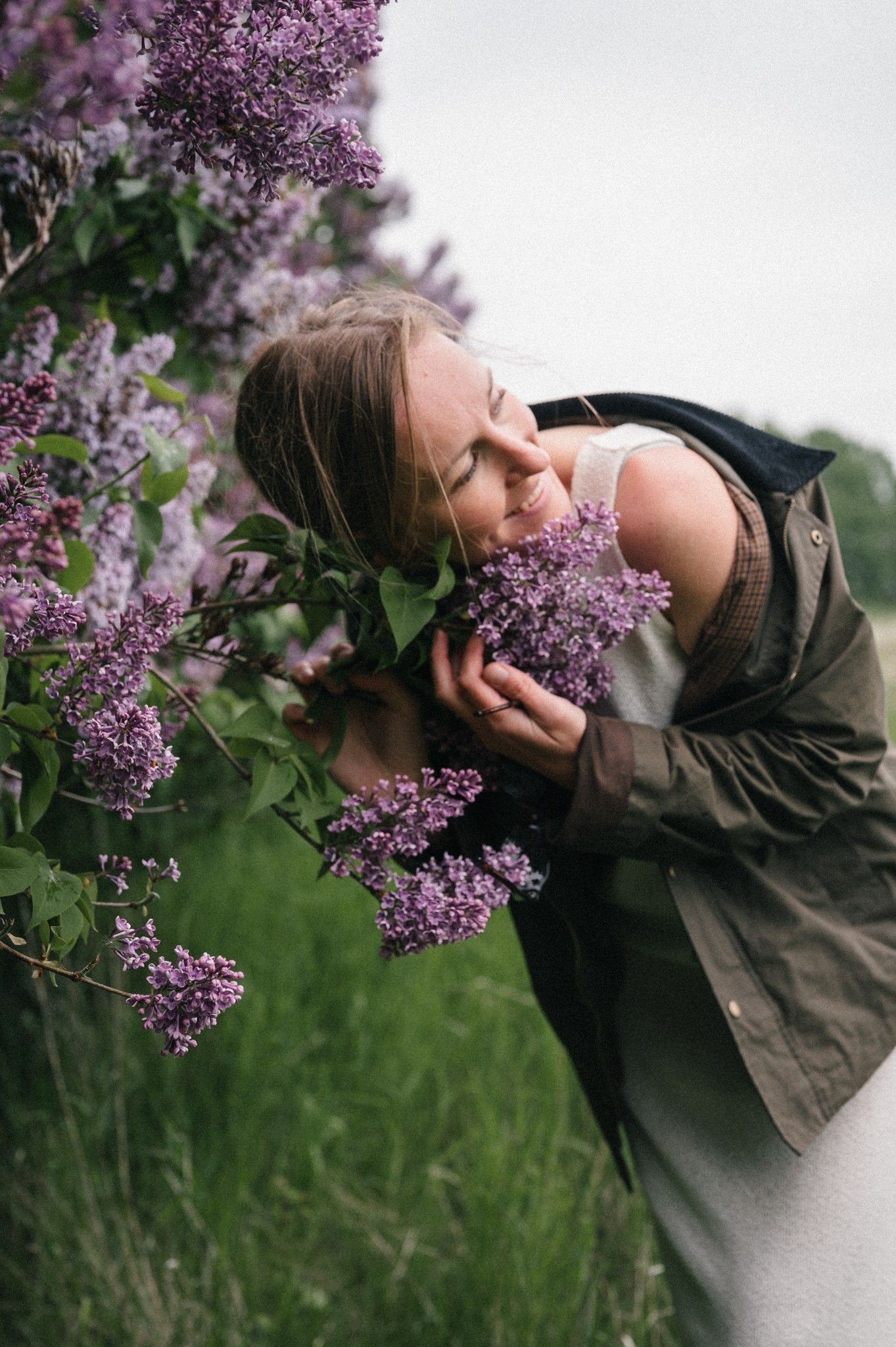 Woman with closed eyes smelling purple lilac flowers outdoors, wearing a green jacket in a lush garden setting.