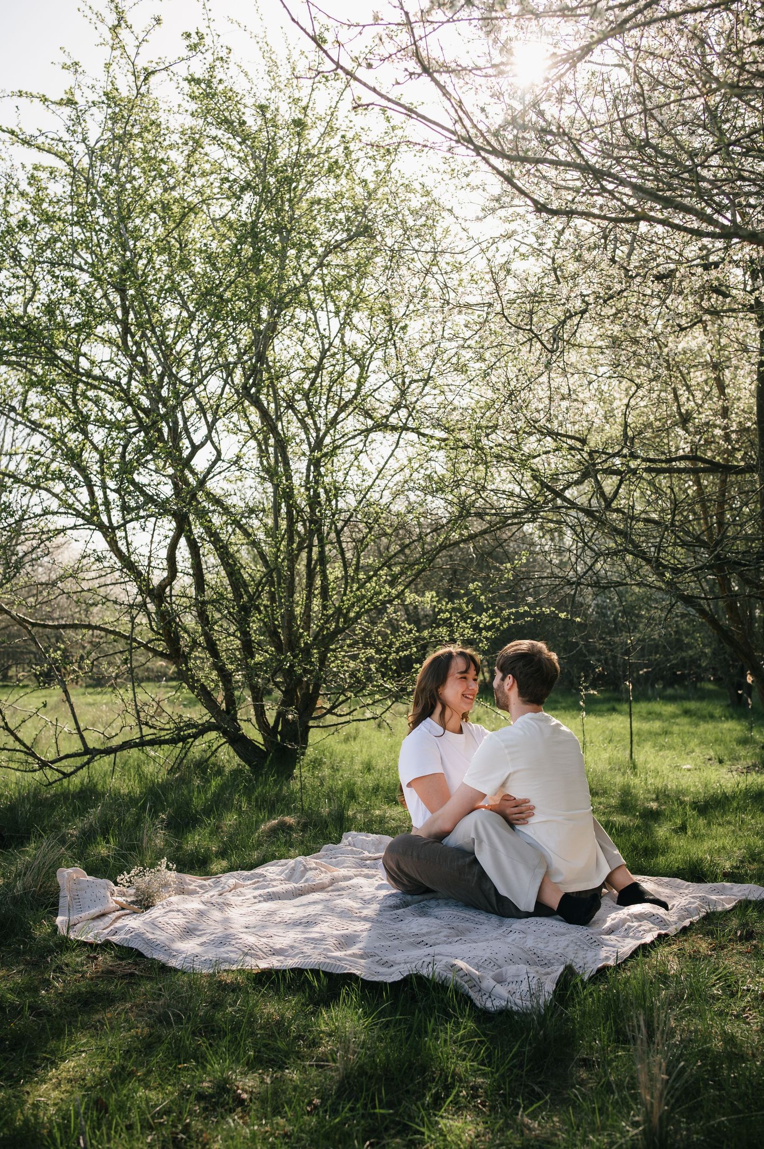 Couple sitting on a blanket in a lush green garden under blooming trees, sharing a romantic moment outdoors.