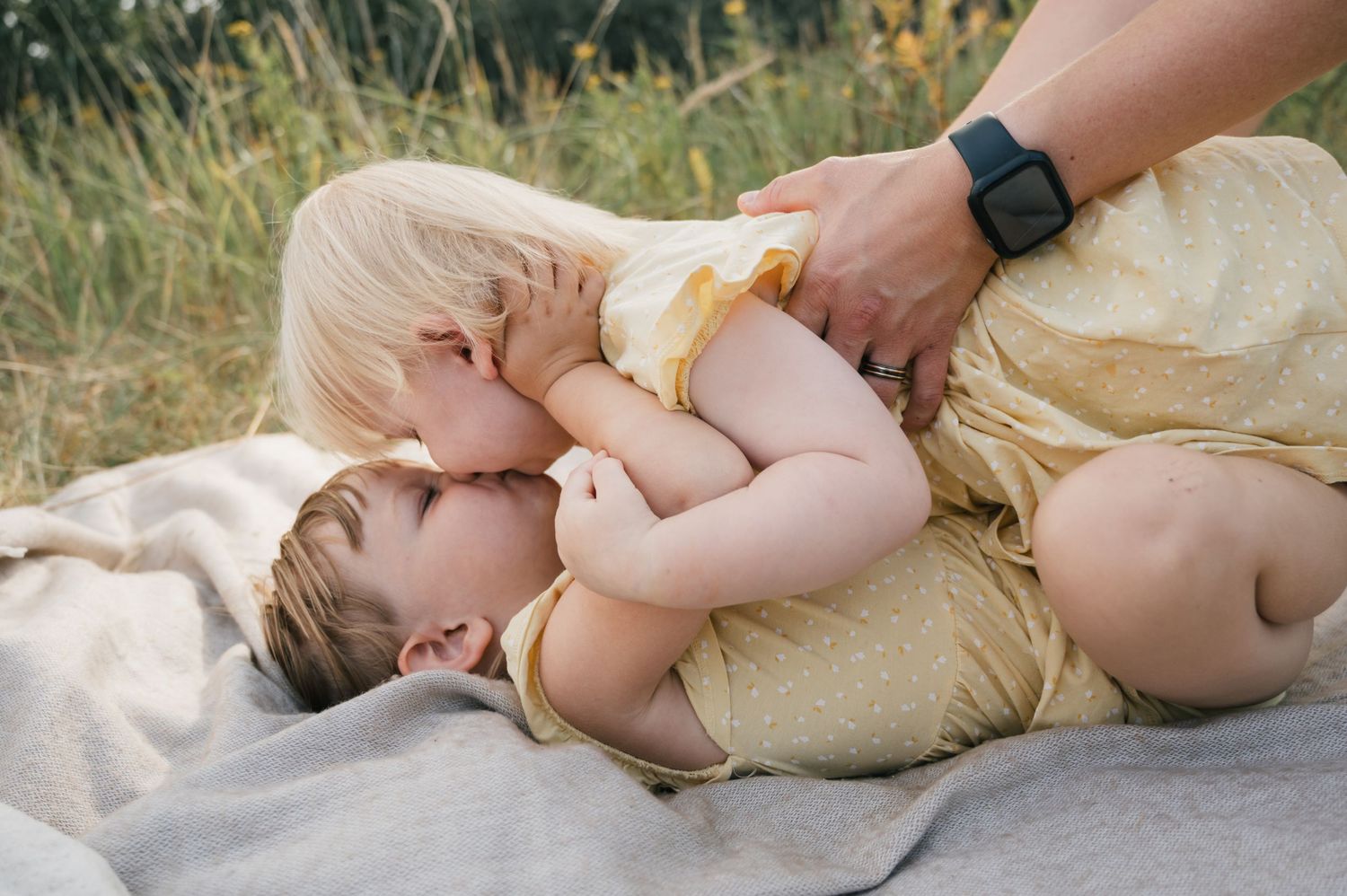 Two young toddlers in yellow outfits cuddle together on a blanket outdoors while an adult watches over them.
