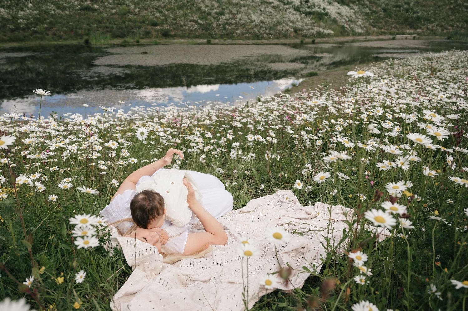 Person lying in a field of white daisies near a reflective lake, wrapped in a lace blanket on a summer day.