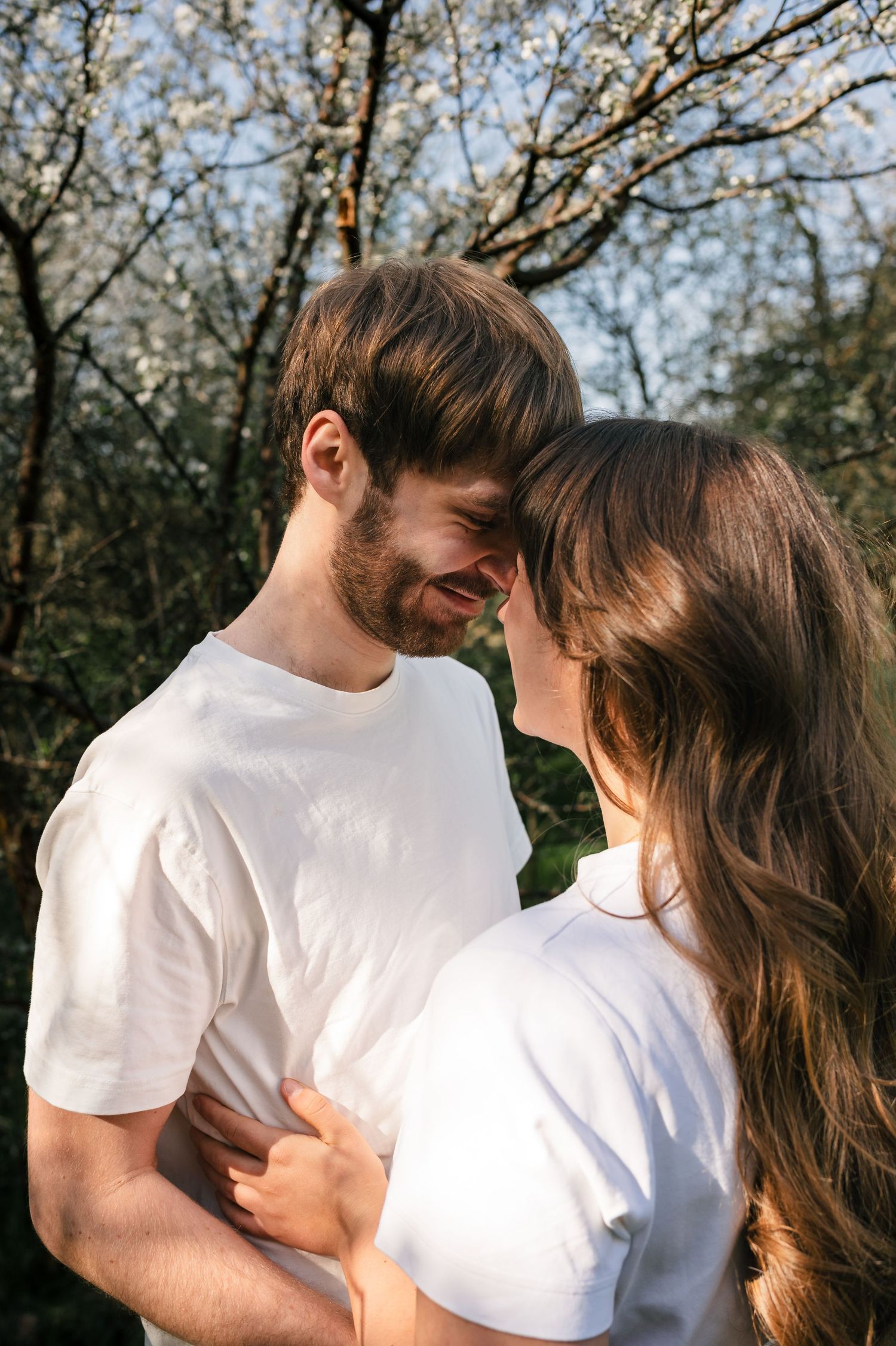 A couple in matching white t-shirts share a tender moment, touching foreheads outdoors near blooming trees.