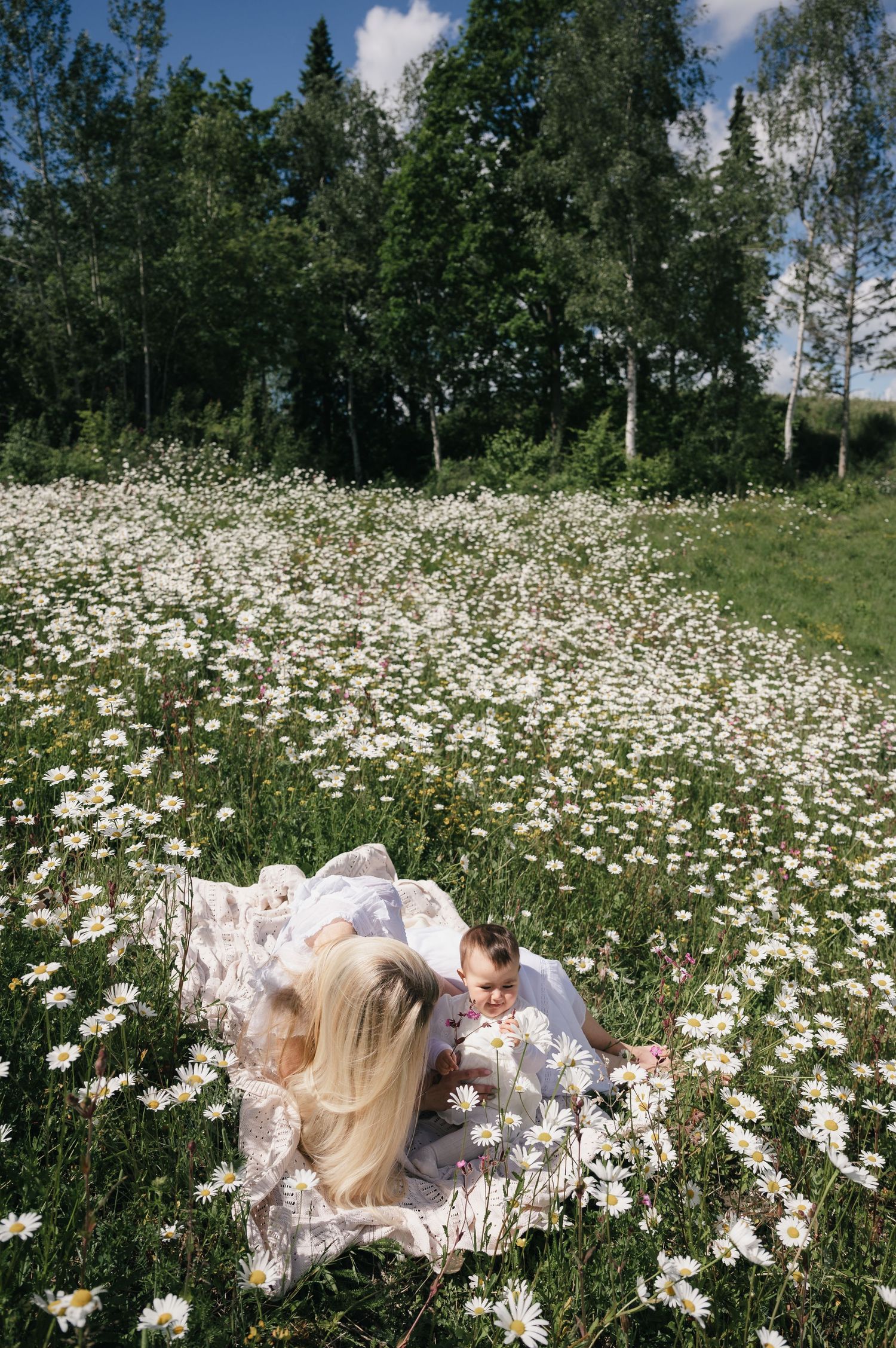 Mother and baby relaxing on a blanket in a sunny daisy field surrounded by green trees.