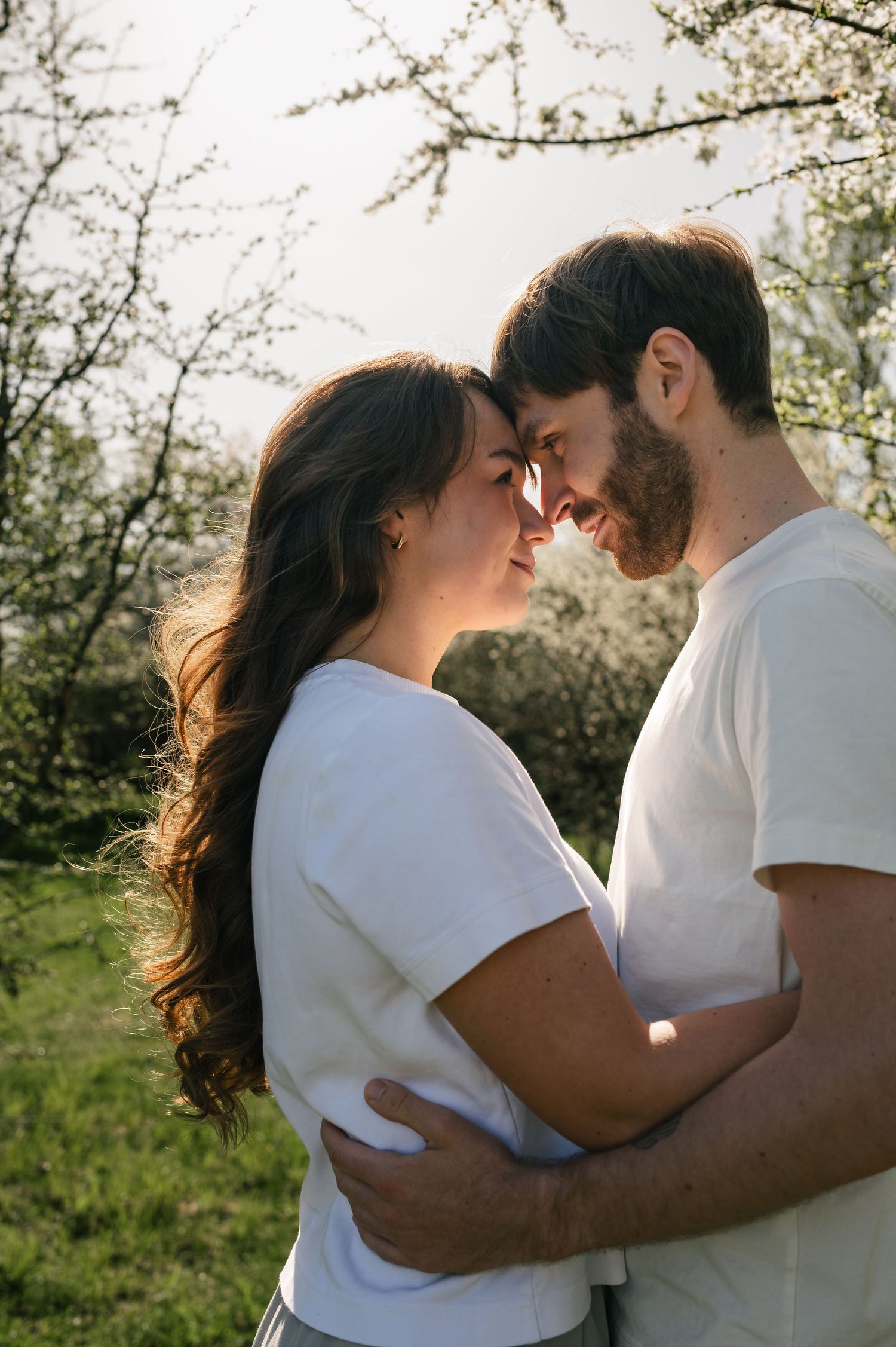 A couple in white t-shirts embrace and touch foreheads in a sunny garden with blooming trees.