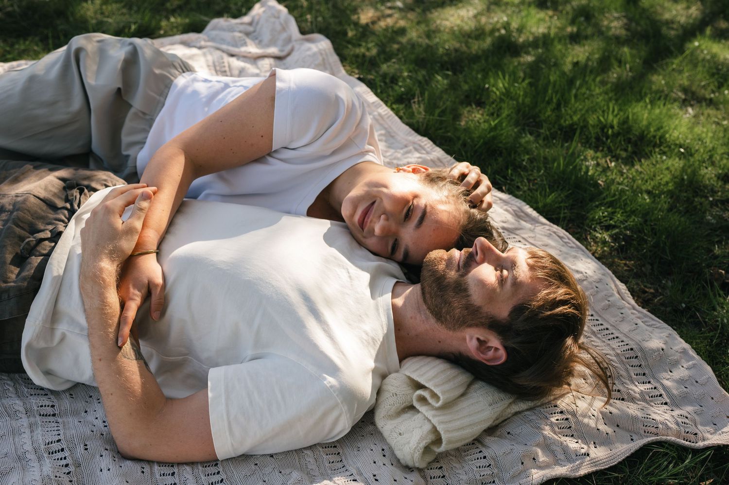 Couple relaxing on a blanket in sunny grass, woman smiling while lying on man's chest outdoors.