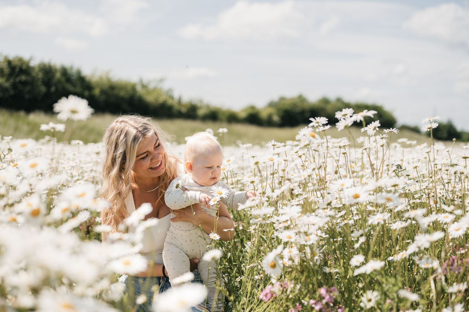 En mor og hendes barn sidder sammen i en blomstrende eng fyldt med hvide margueritter på en solrig sommerdag.