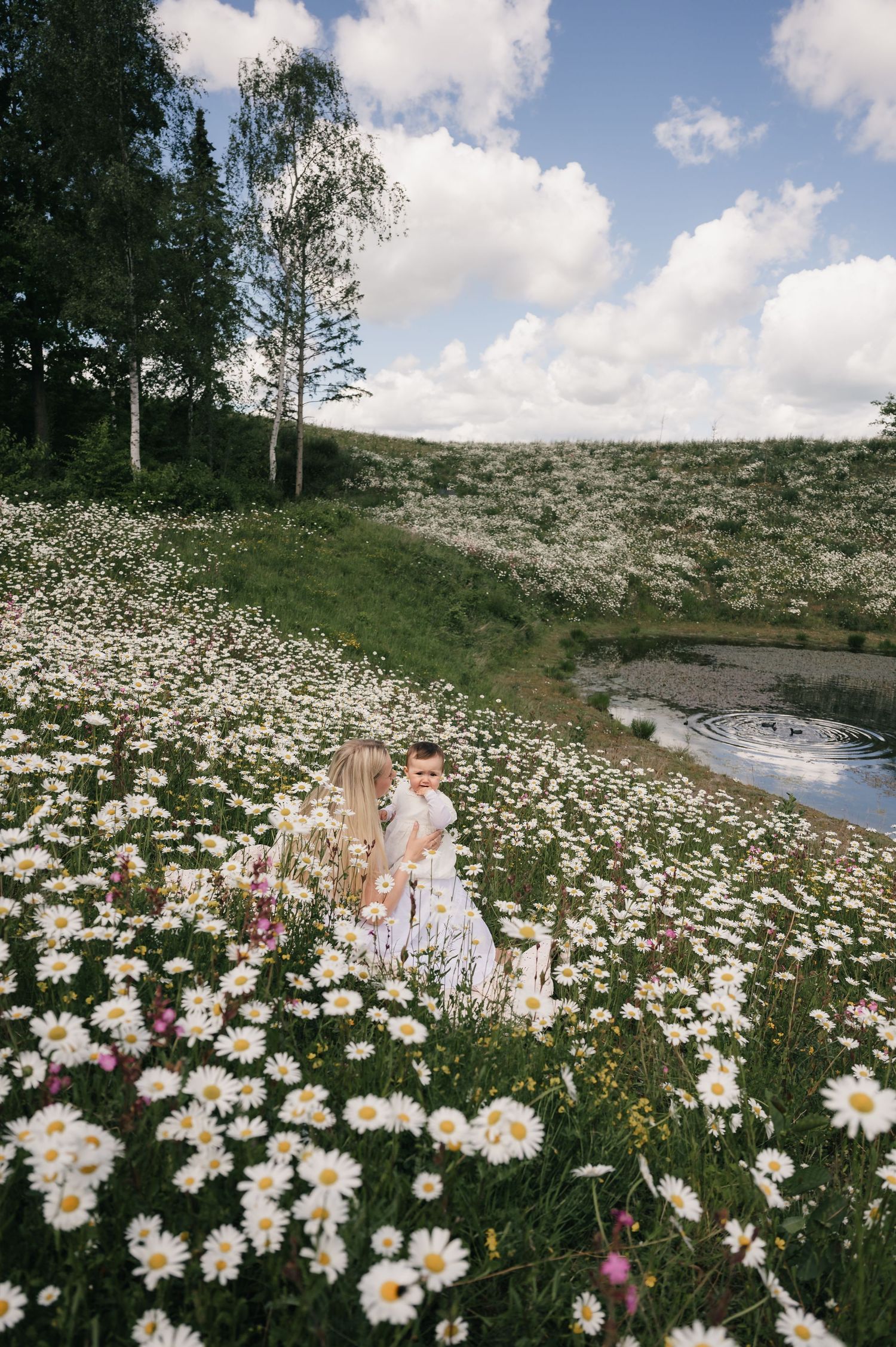 Woman and child sitting in a vast daisy field near a pond, surrounded by lush trees under a partly cloudy sky.
