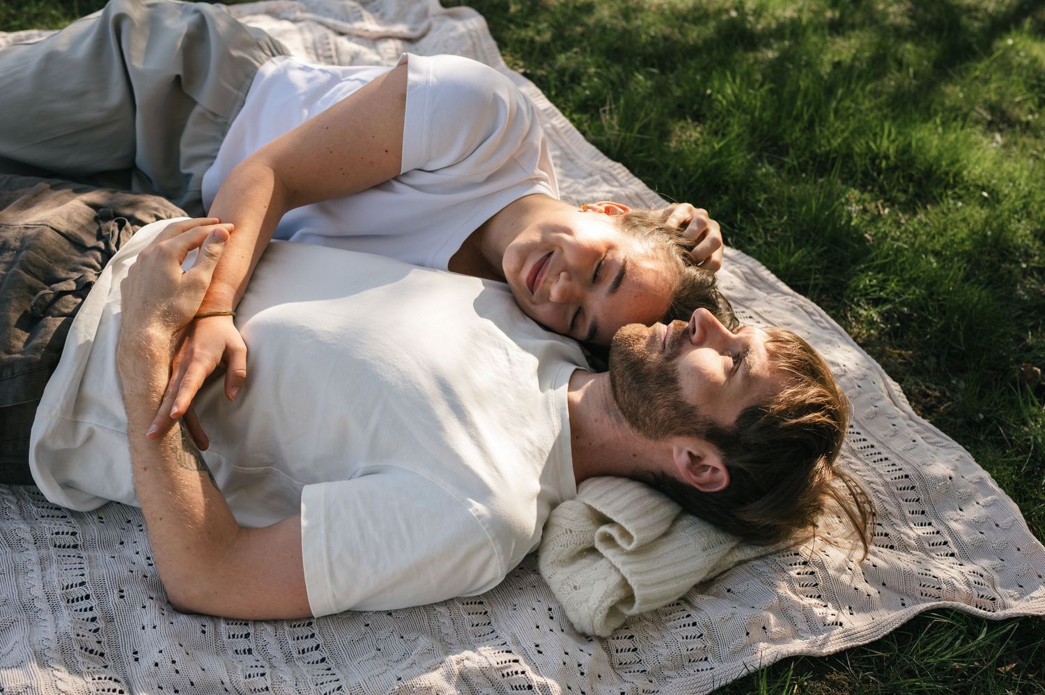 Couple lying on a blanket in the grass, smiling and embracing on a sunny day outdoors.