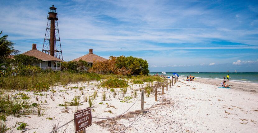 Historic lighthouse and cottages rise above a white sandy beach path with sea oats and turquoise Gulf waters under a blue sky.