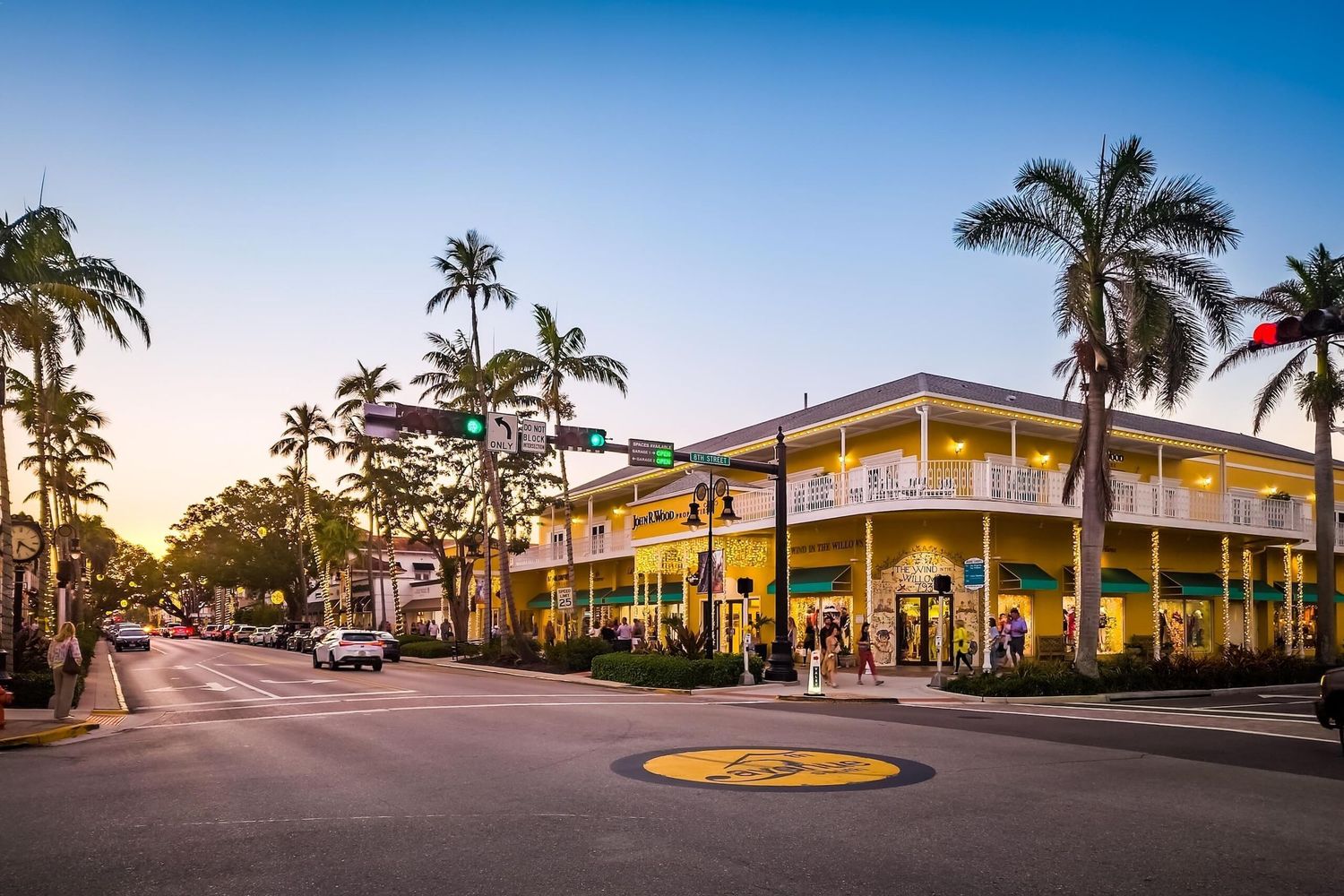 Palm trees line a quiet downtown street at dusk, with a two-story shopping building, traffic lights and a yellow medallion in the road.