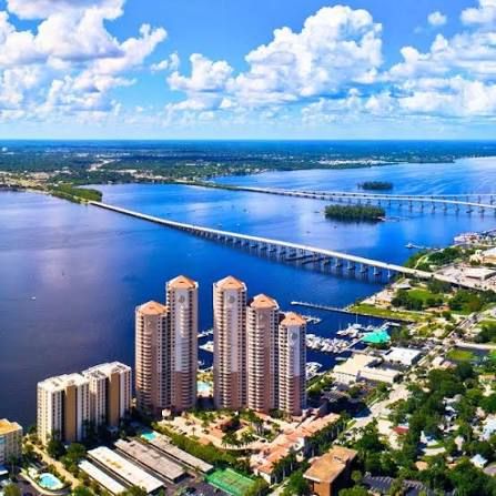 Aerial view of waterfront high-rise condo towers beside a wide river with two long bridges, marinas, and green shoreline under a blue sky.