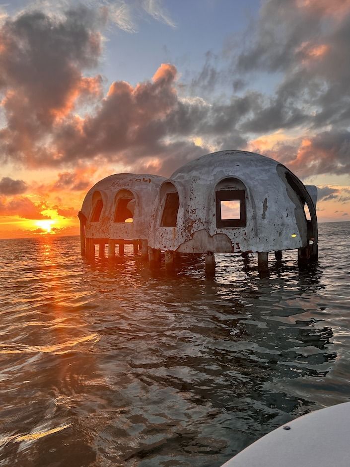Weathered dome-shaped houses on stilts stand in calm ocean water at sunset, with orange and pink clouds glowing across the sky.