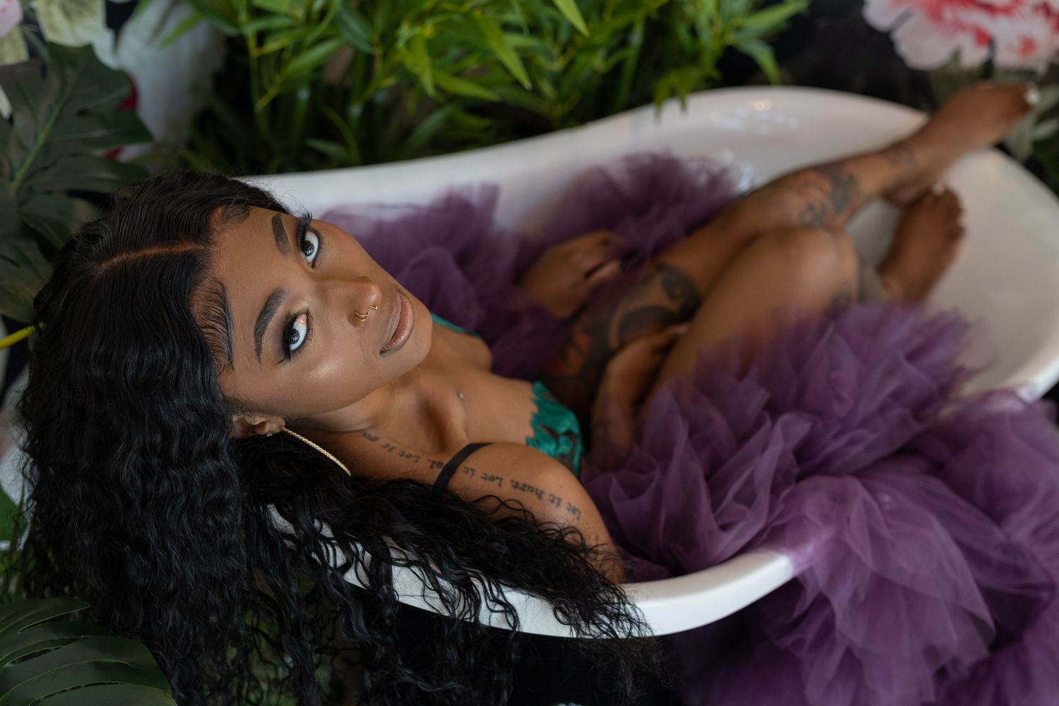 A reclining person in purple tulle fabric lays in a clawfoot bathtub surrounded by greenery.