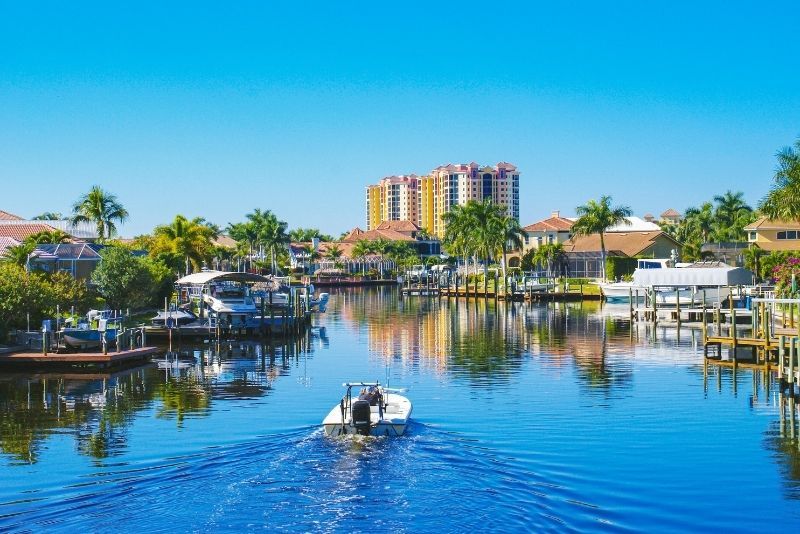 Small motorboat cruising a sunny waterfront canal lined with docks, palm trees, and waterfront homes with a high-rise condo in the distance.