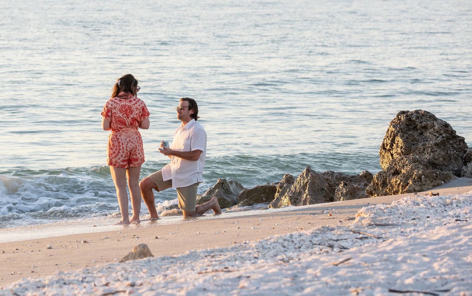 A romantic beachside marriage proposal scene at sunset with ocean waves and rocks in the background.
