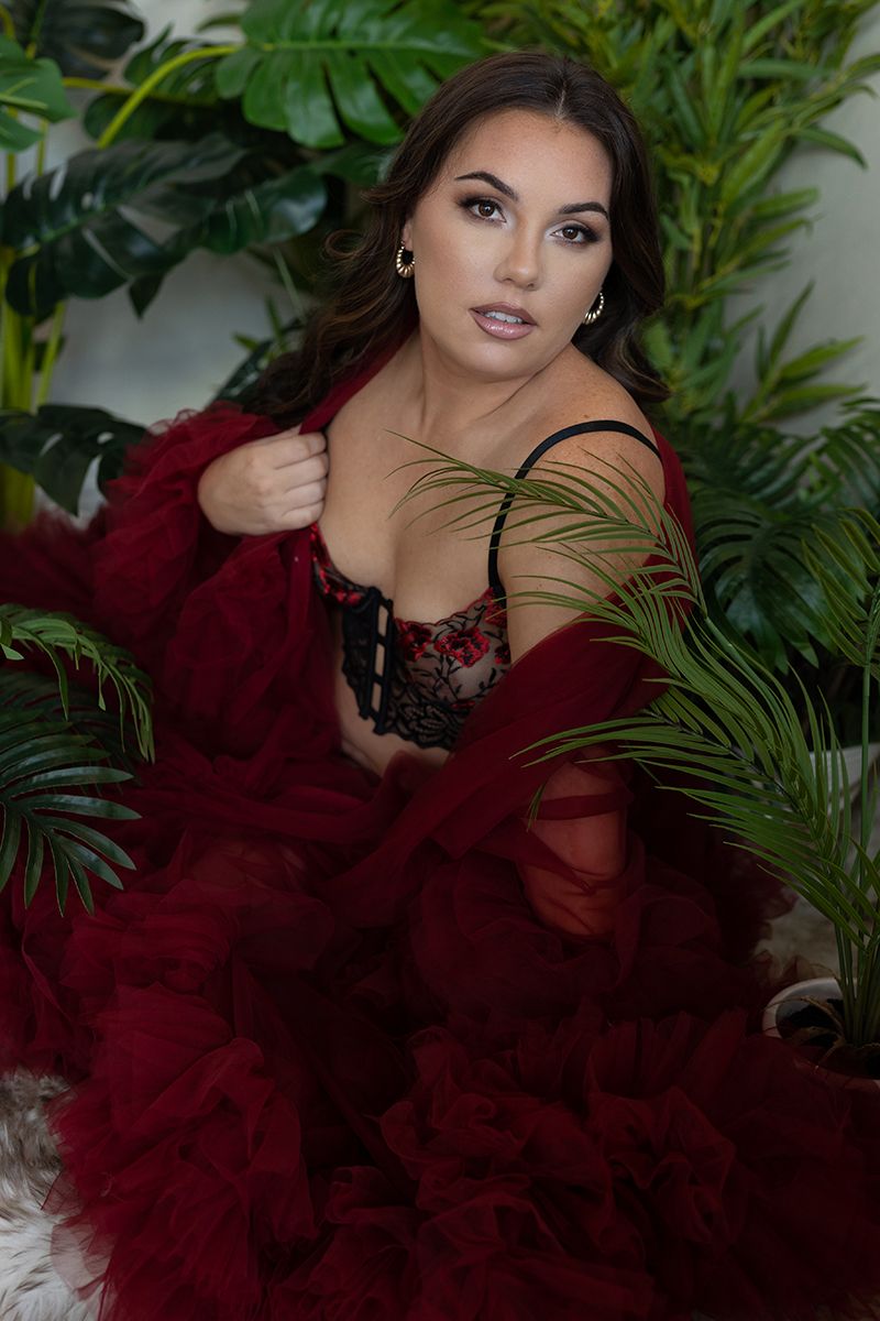 Glamour portrait surrounded by tropical plants featuring burgundy robe and lace against green foliage background.