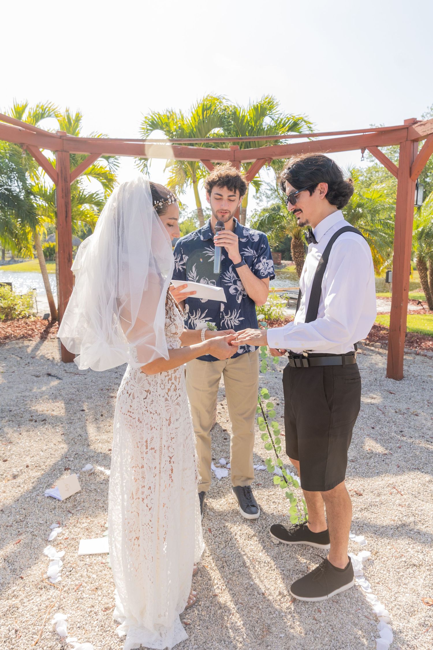 A wedding ceremony takes place outdoors under a wooden pergola with palm trees in the background on a sunny day.