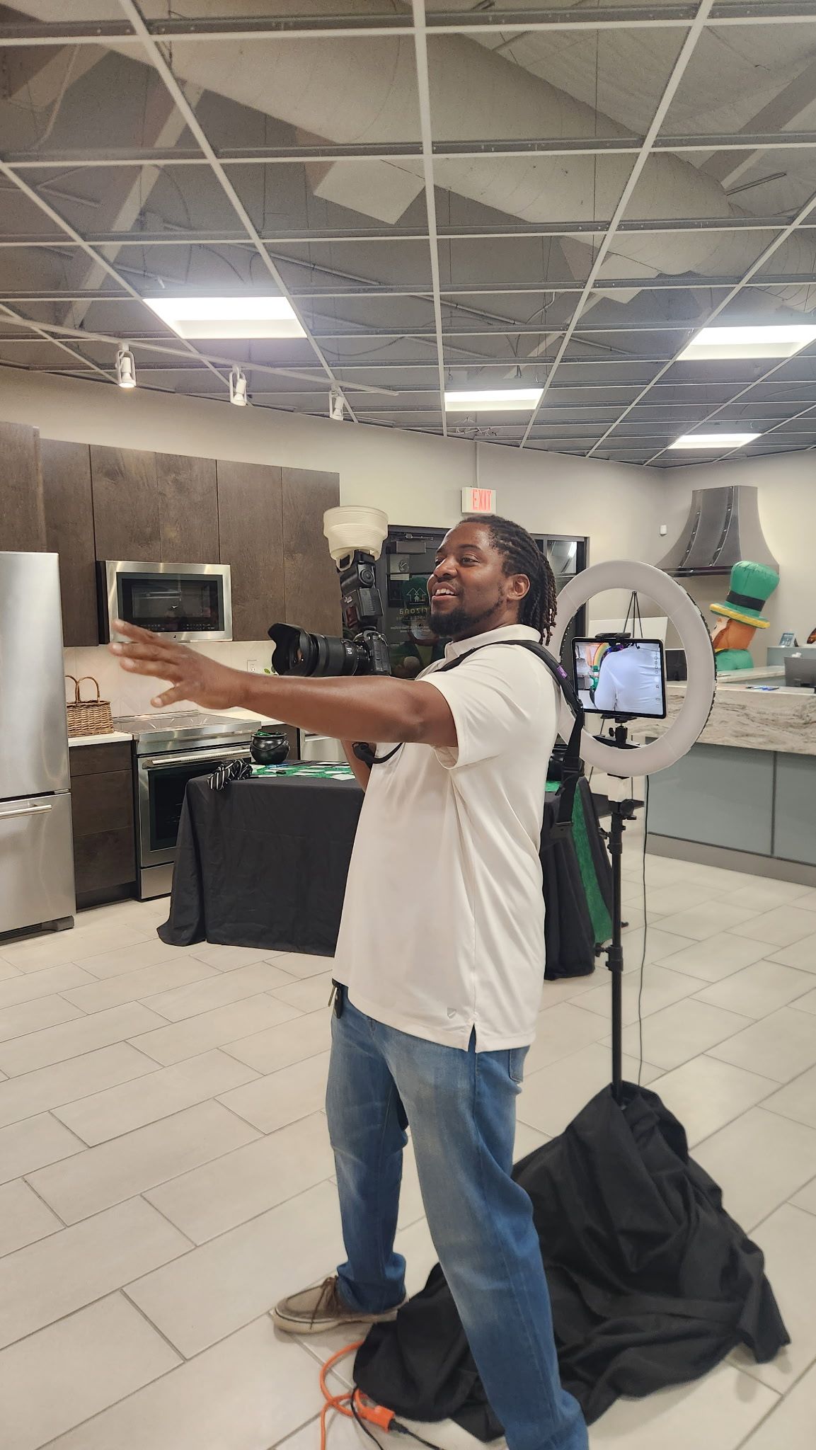 A smiling photographer with dreadlocks gestures while holding a camera near a ring light in a kitchen studio.