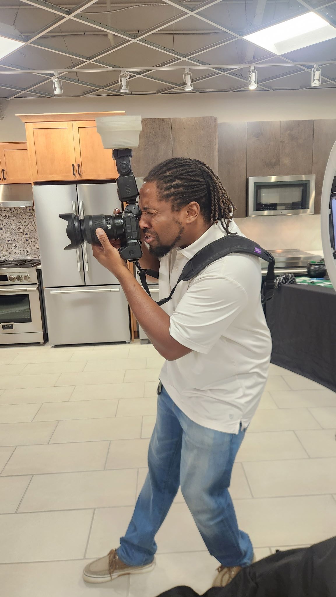 A photographer with dreadlocks shoots with a DSLR camera in a modern kitchen showroom.