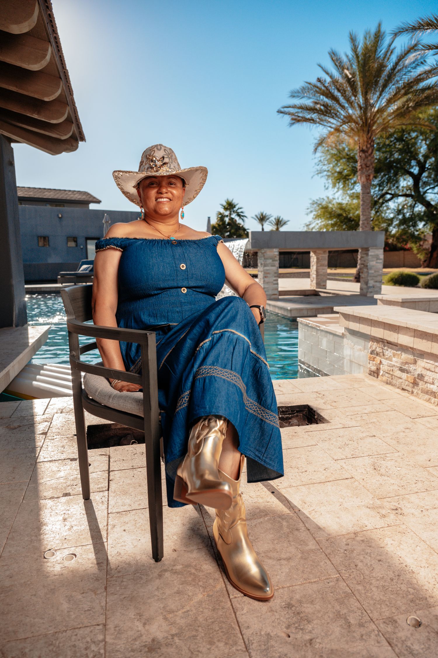Woman in denim dress, cowboy hat, and gold boots sitting poolside with palm trees in sunny backyard.
