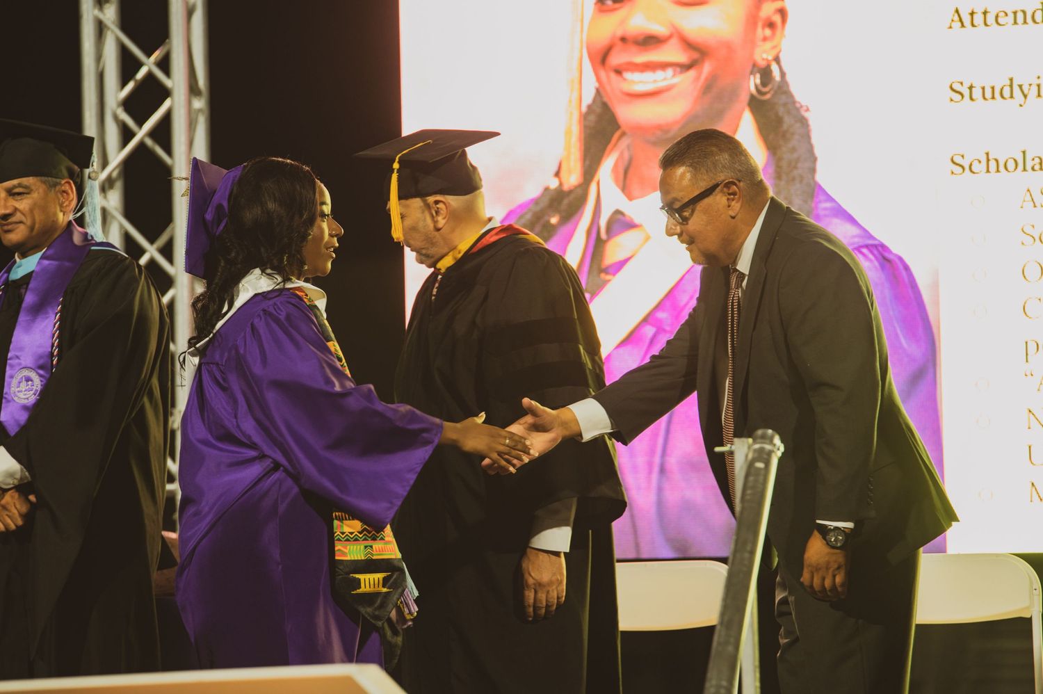 Graduates in purple robes shake hands with faculty during a university commencement ceremony on stage.