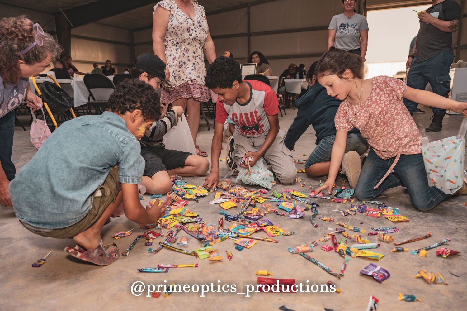 Children and adults gather on the floor sorting colorful candies at an outdoor event organized by Prime Optics Productions.