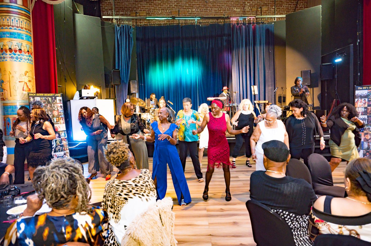 Diverse group of people dancing and socializing at a vibrant indoor party event with teal curtains and colorful decor.