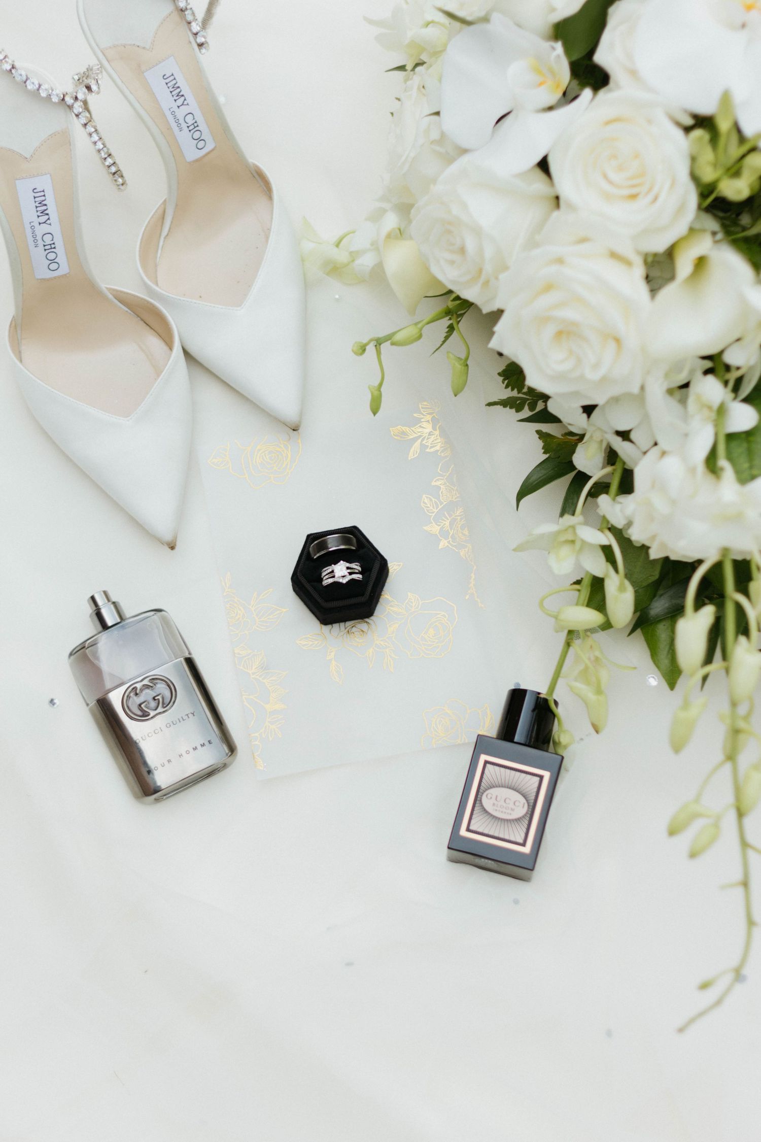 Elegant wedding accessories including white roses, perfume bottles, and jewelry arranged on white background.