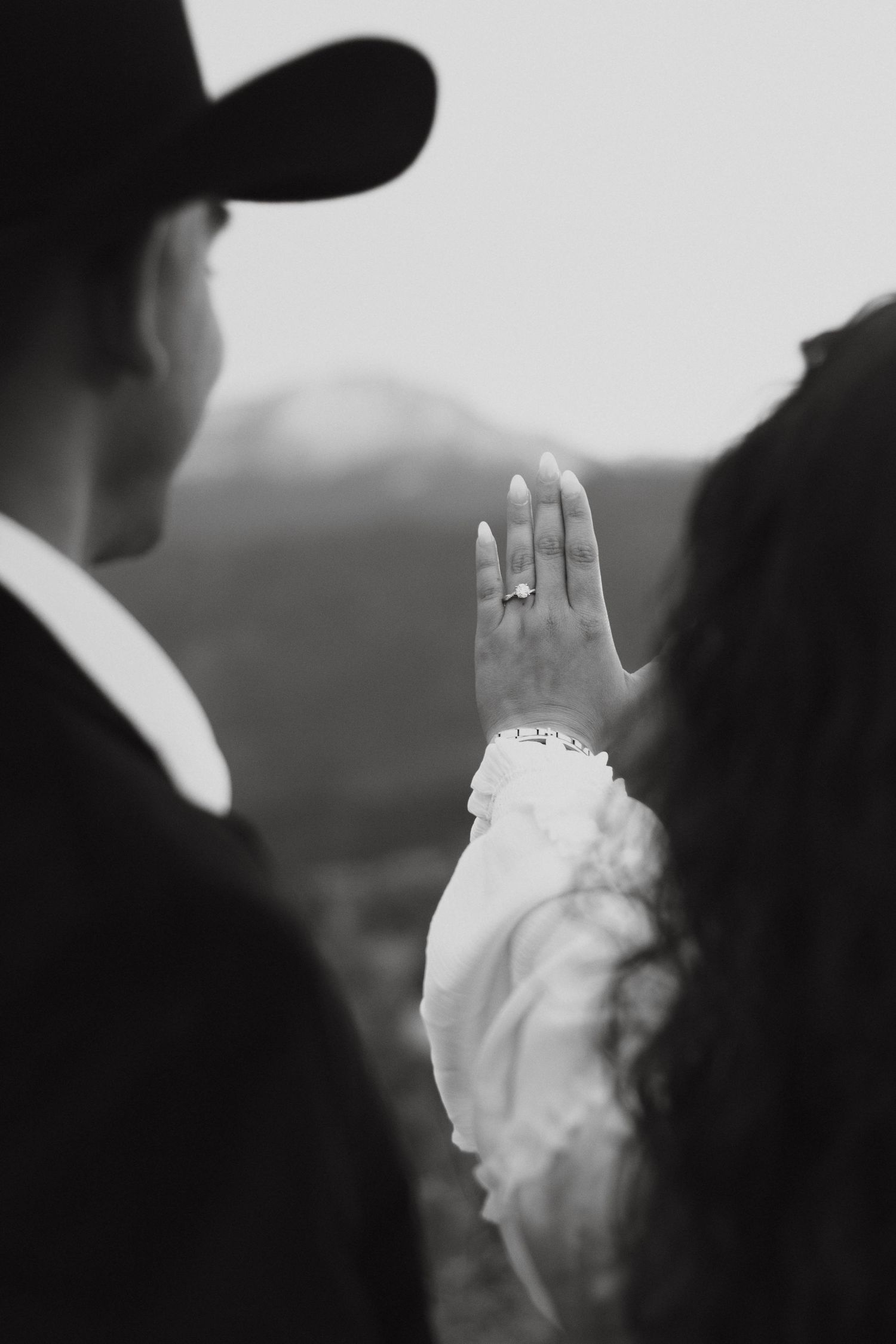 A close-up black and white photo showing an engagement ring on a raised hand against a blurred outdoor background.