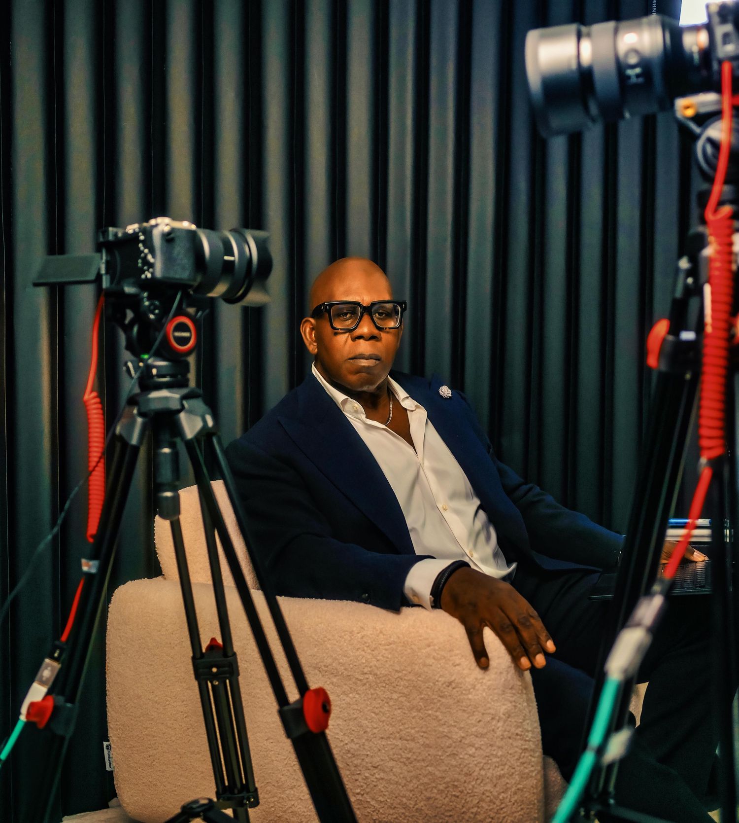 A bald man in a navy blazer sits in a studio chair surrounded by professional cameras on tripods.