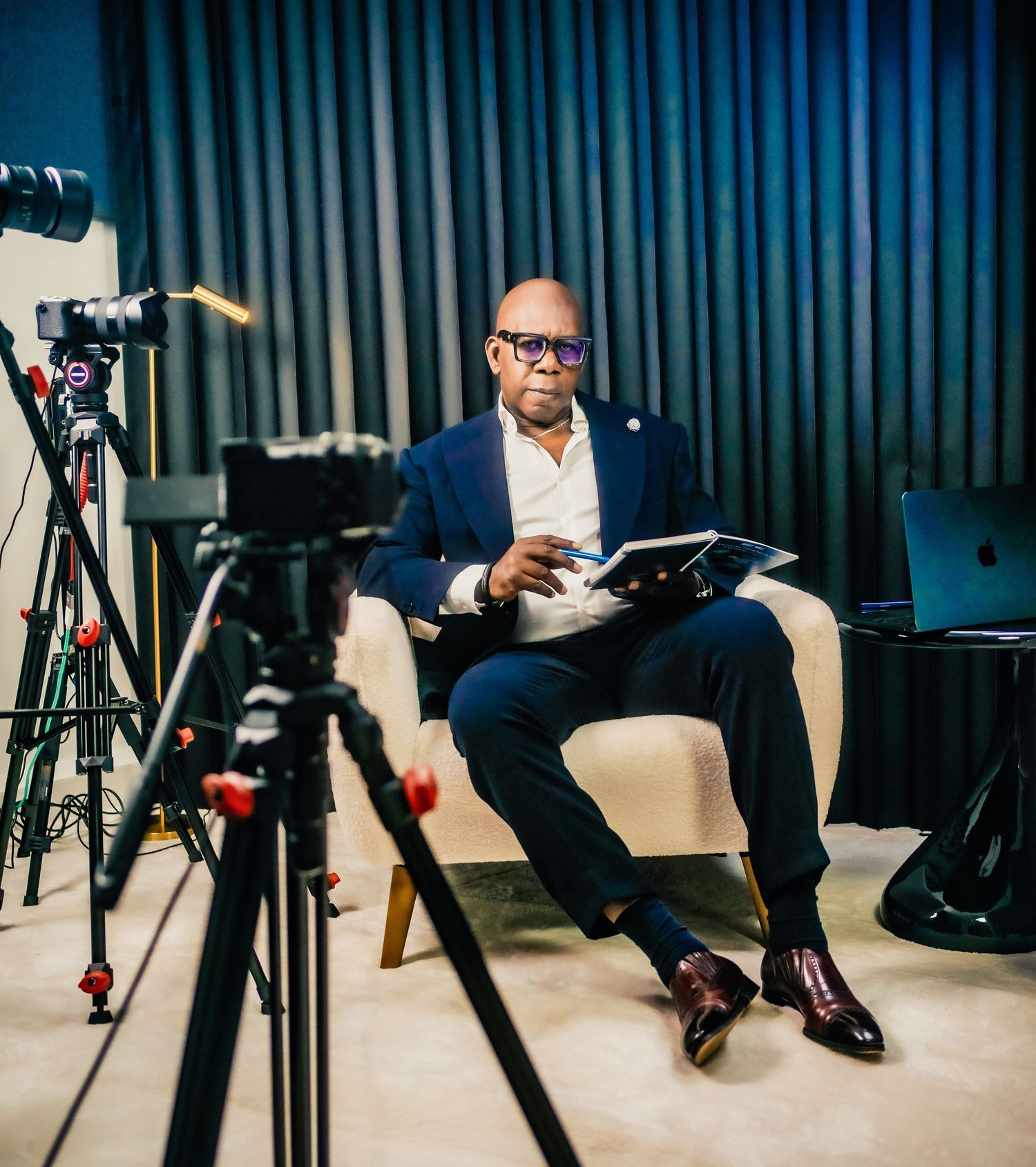 Professional man in navy suit reads documents while seated between cameras in a studio setting.