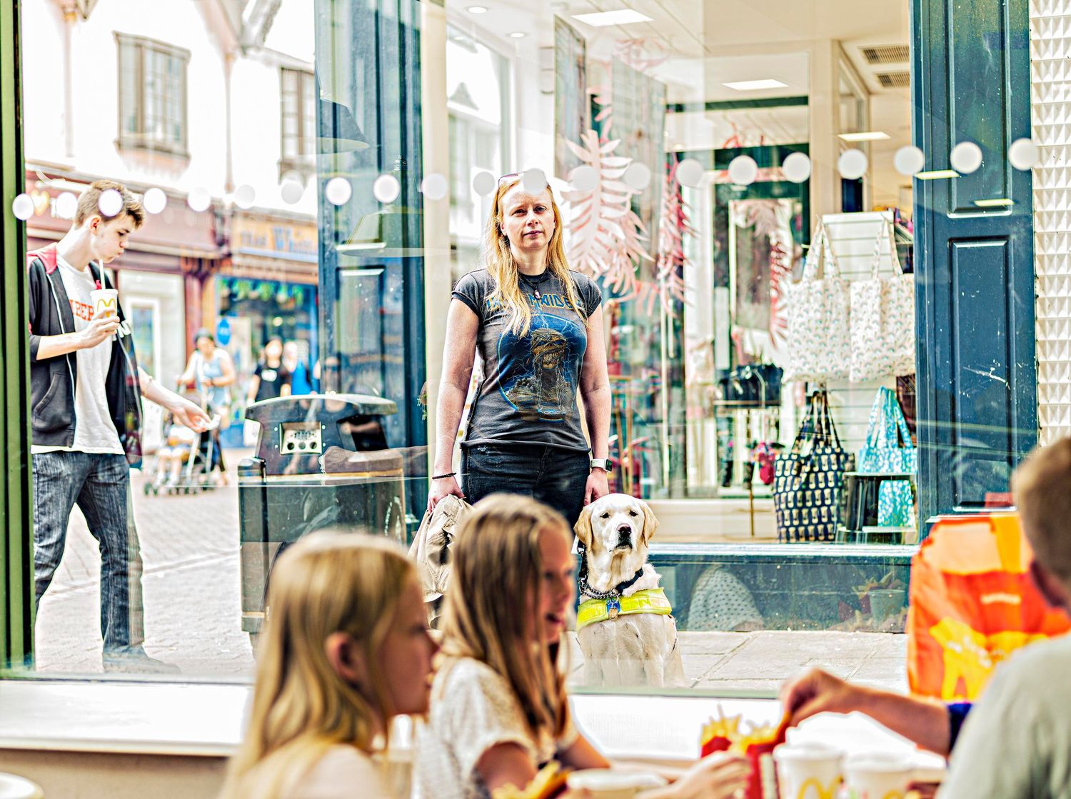 Woman walking a golden retriever guide dog through a busy shopping street past storefronts.