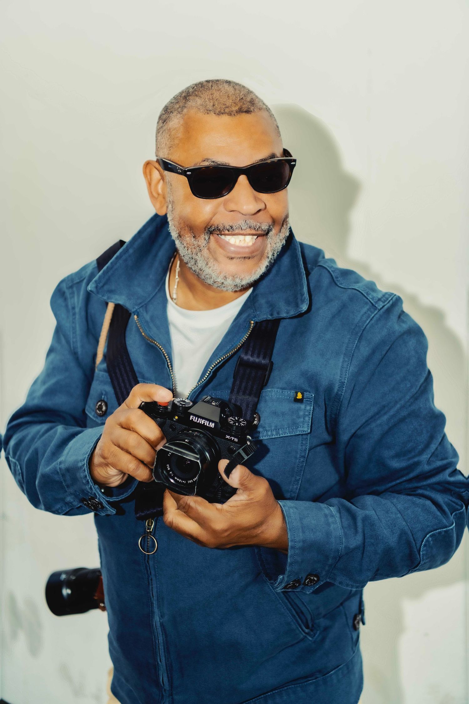 Smiling photographer wearing sunglasses and blue denim jacket holding a professional camera against white background.