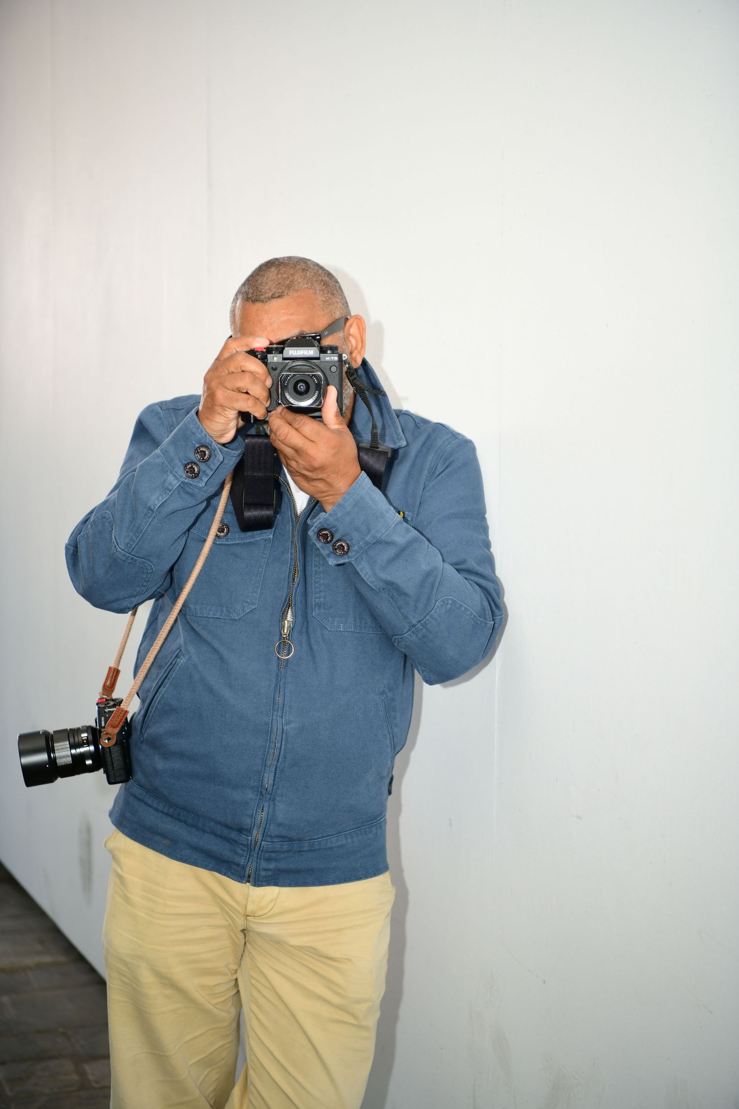 A man in a blue jacket and khaki pants holds a vintage film camera up to his face while another camera hangs at his side.