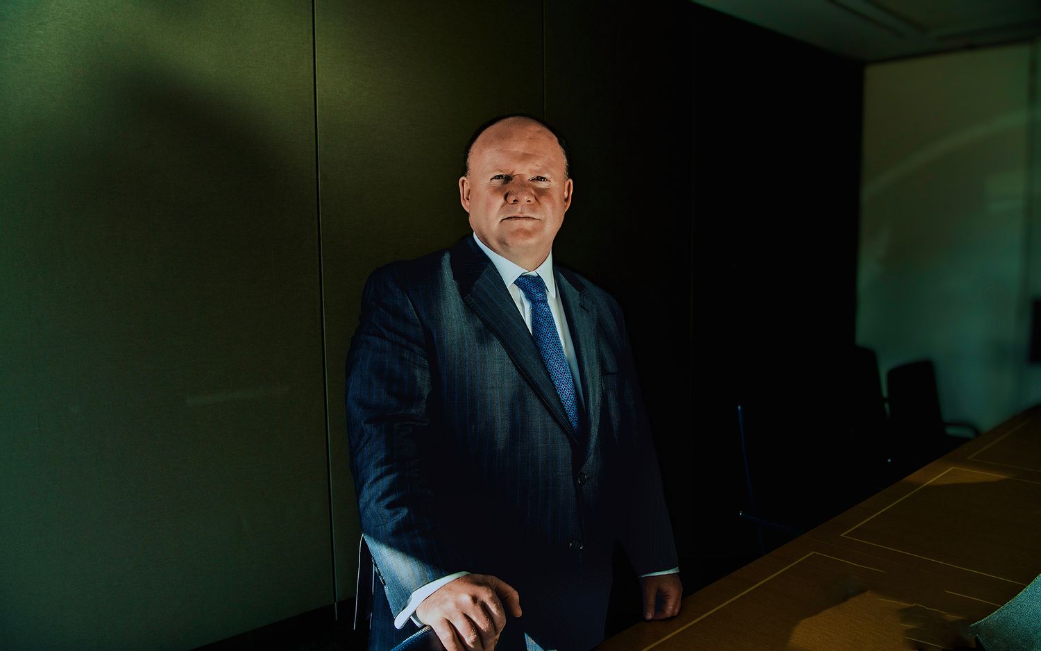 Professional businessman in navy pinstripe suit standing confidently in modern boardroom with dark walls.