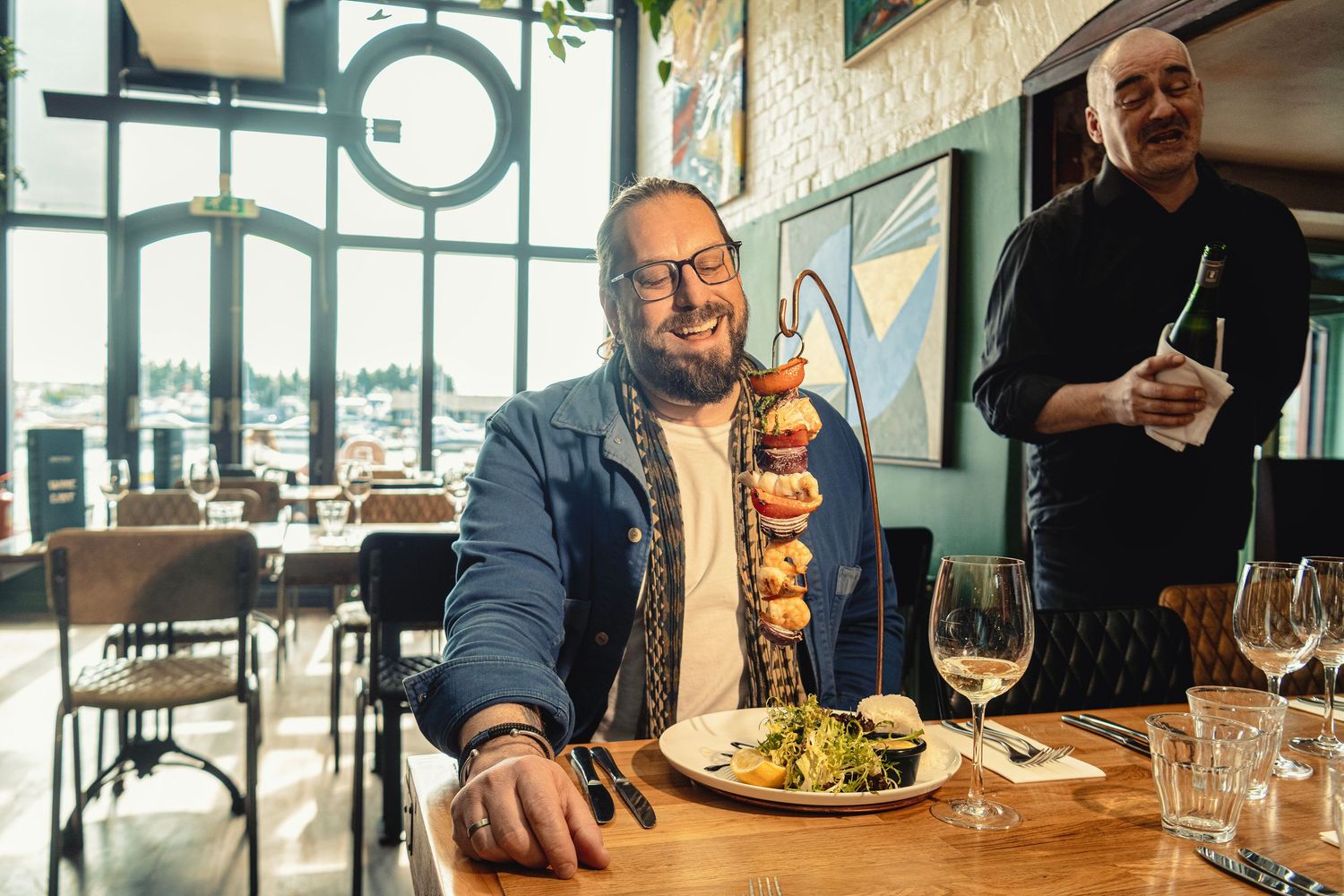 Man smiling at restaurant table with colorful food skewer on plate, waiter standing in background.