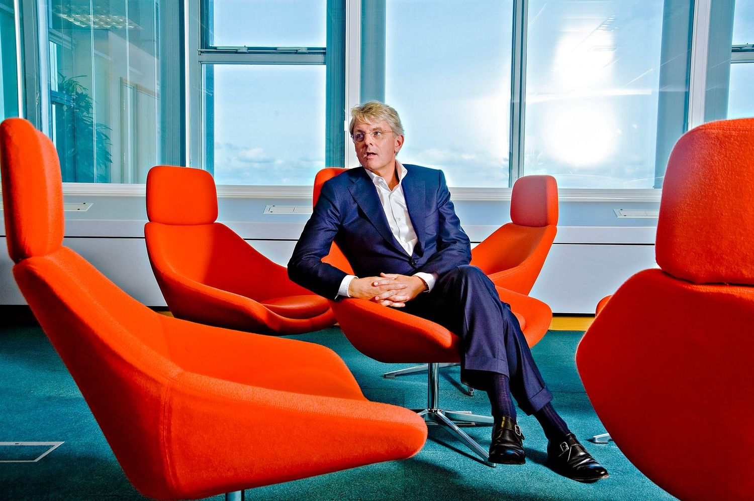 Man in blue suit sitting among vibrant orange modern chairs in a bright contemporary office space.