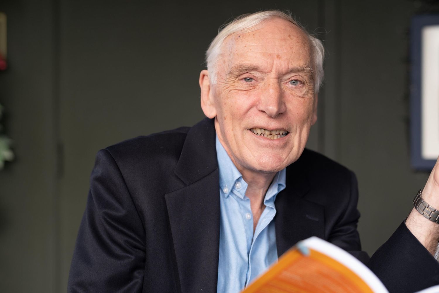 Elderly man in dark blazer and blue shirt smiling while holding an open book indoors.