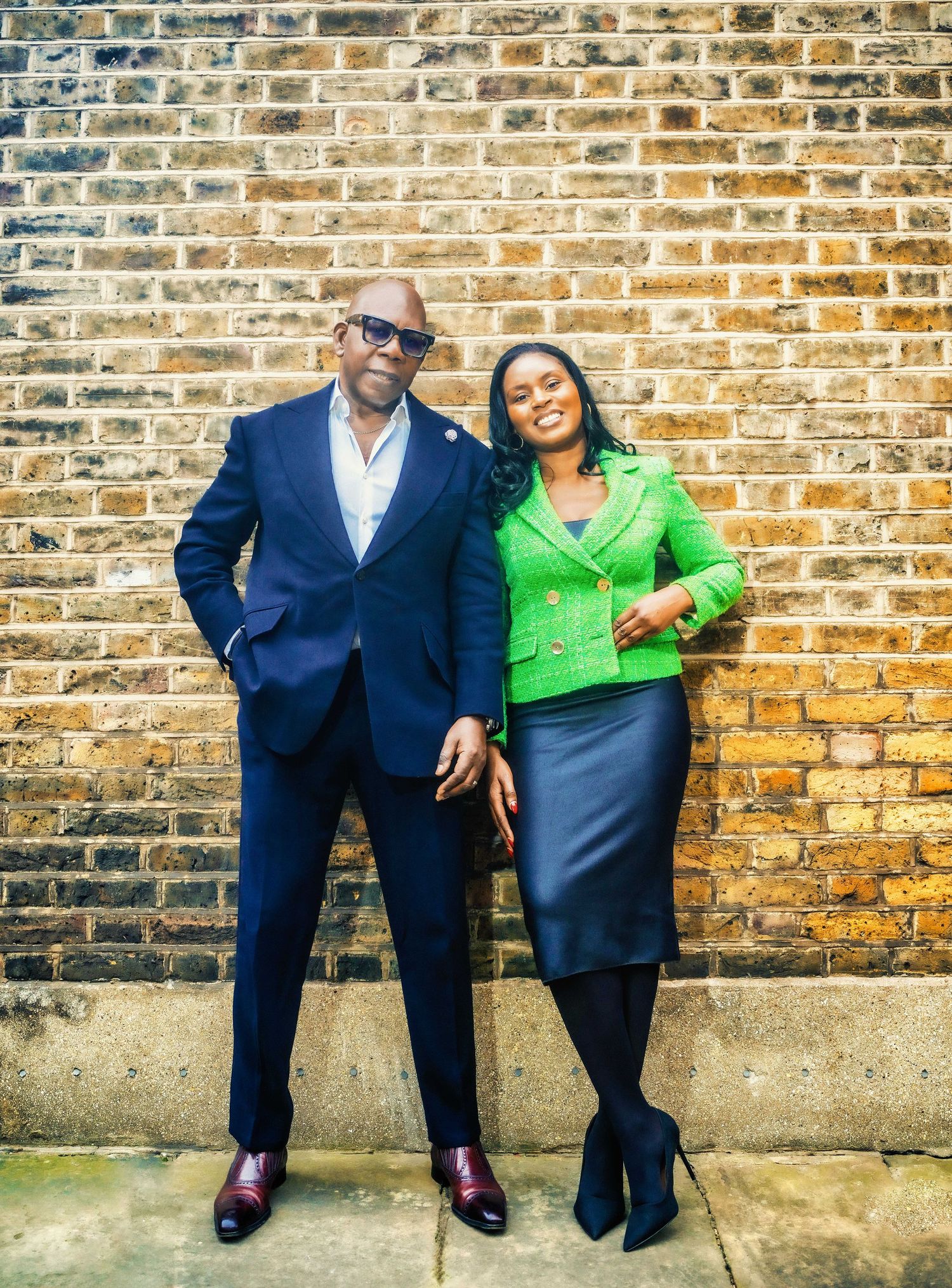 A man in a navy suit and a woman in a green jacket and gray skirt pose together against a brick wall.